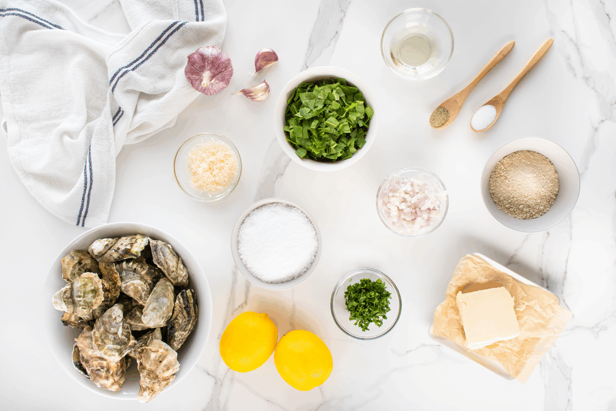 Overhead view of ingredients on a marble surface including oysters, two lemons, butter, chopped herbs, grated cheese, minced shallots, breadcrumbs, garlic, a bowl of salt, and small bowls with seasonings and oil.