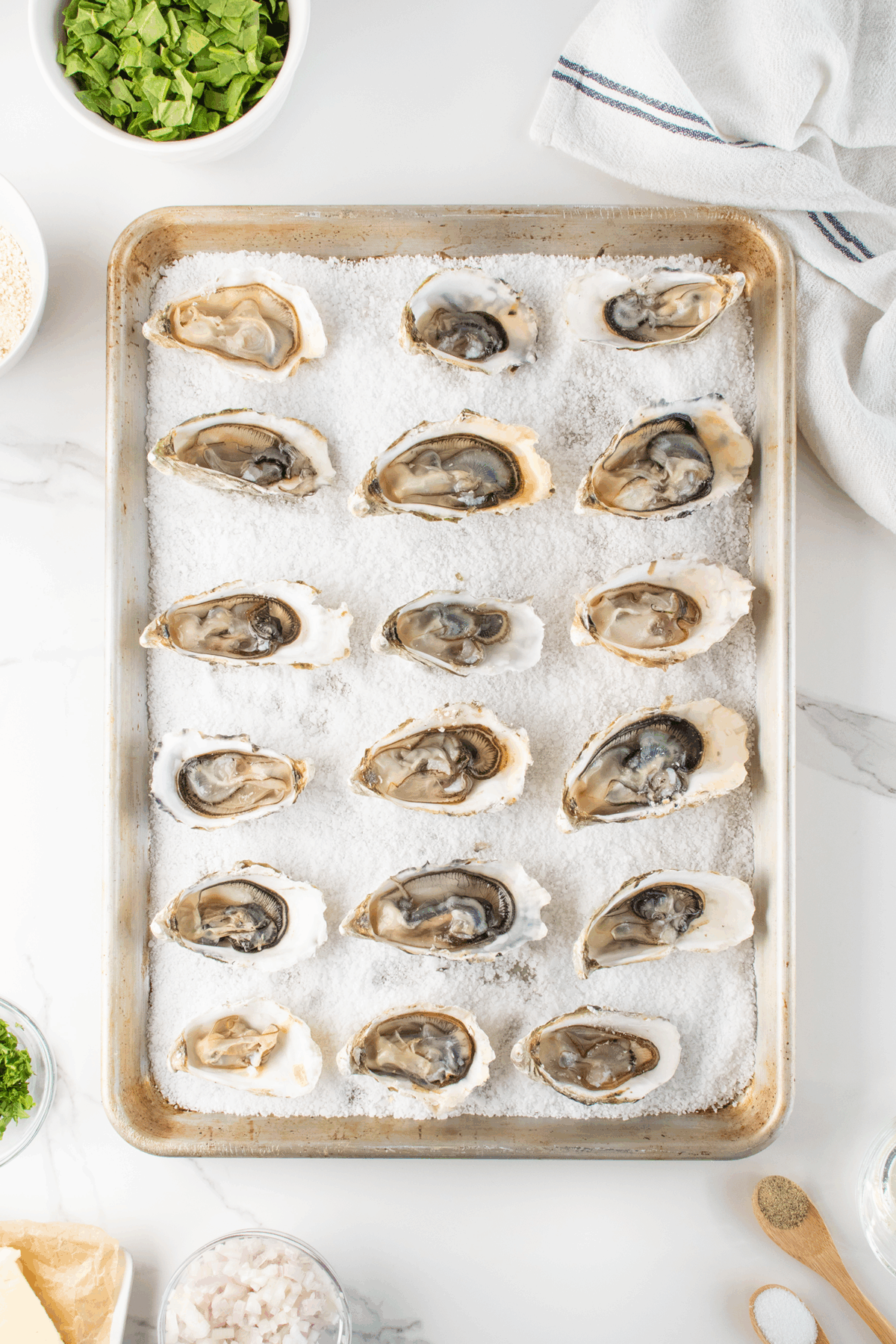 A baking sheet lined with coarse salt holds eighteen opened oysters on the half shell, ready for cooking. Surrounding the pan are bowls of chopped greens, breadcrumbs, herbs, and butter on a marble countertop.