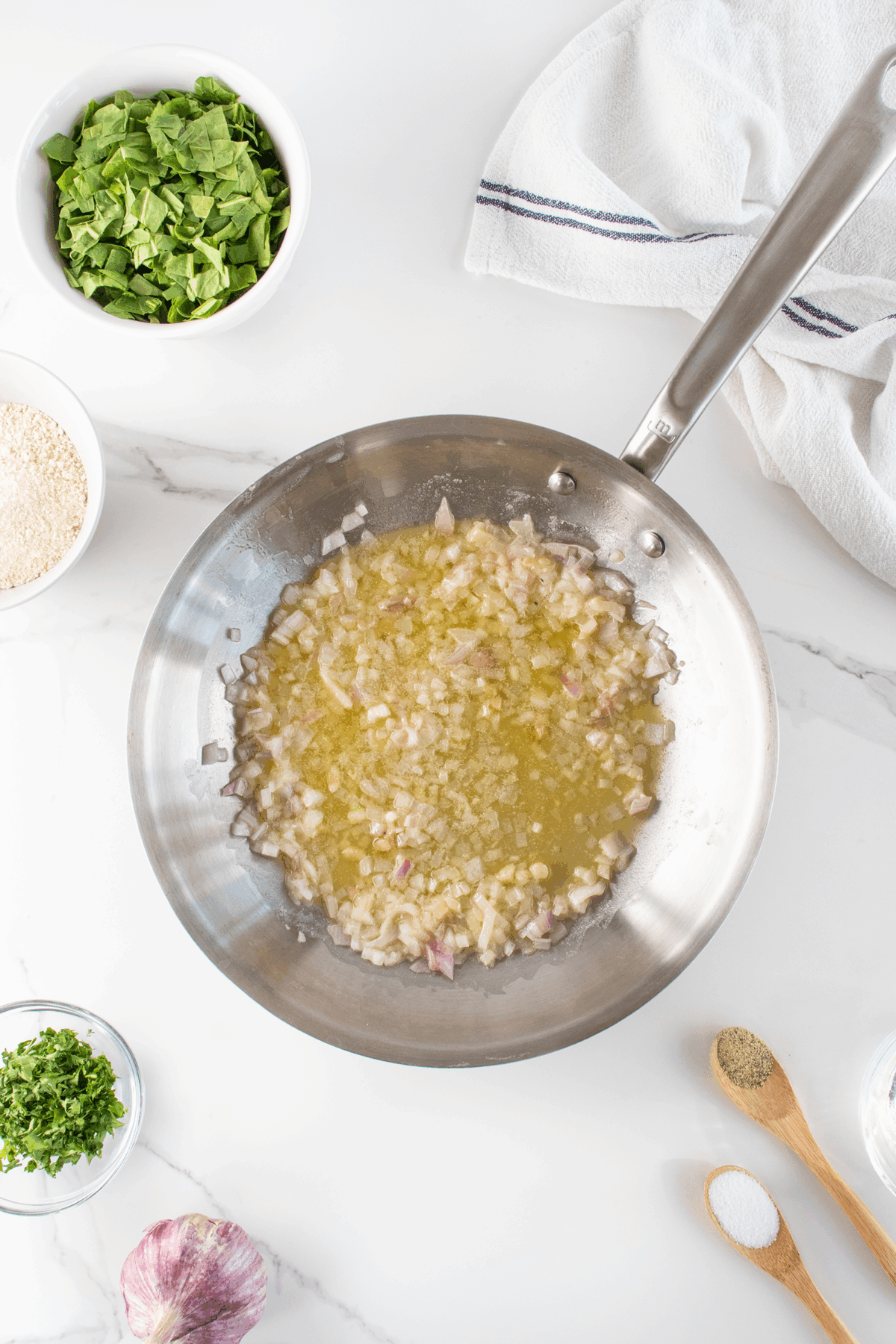 A stainless steel pan with chopped onions sautéing in oil, surrounded by bowls of chopped spinach, parsley, uncooked rice, a garlic bulb, a spoon of salt, and a spoon of black pepper on a white surface.