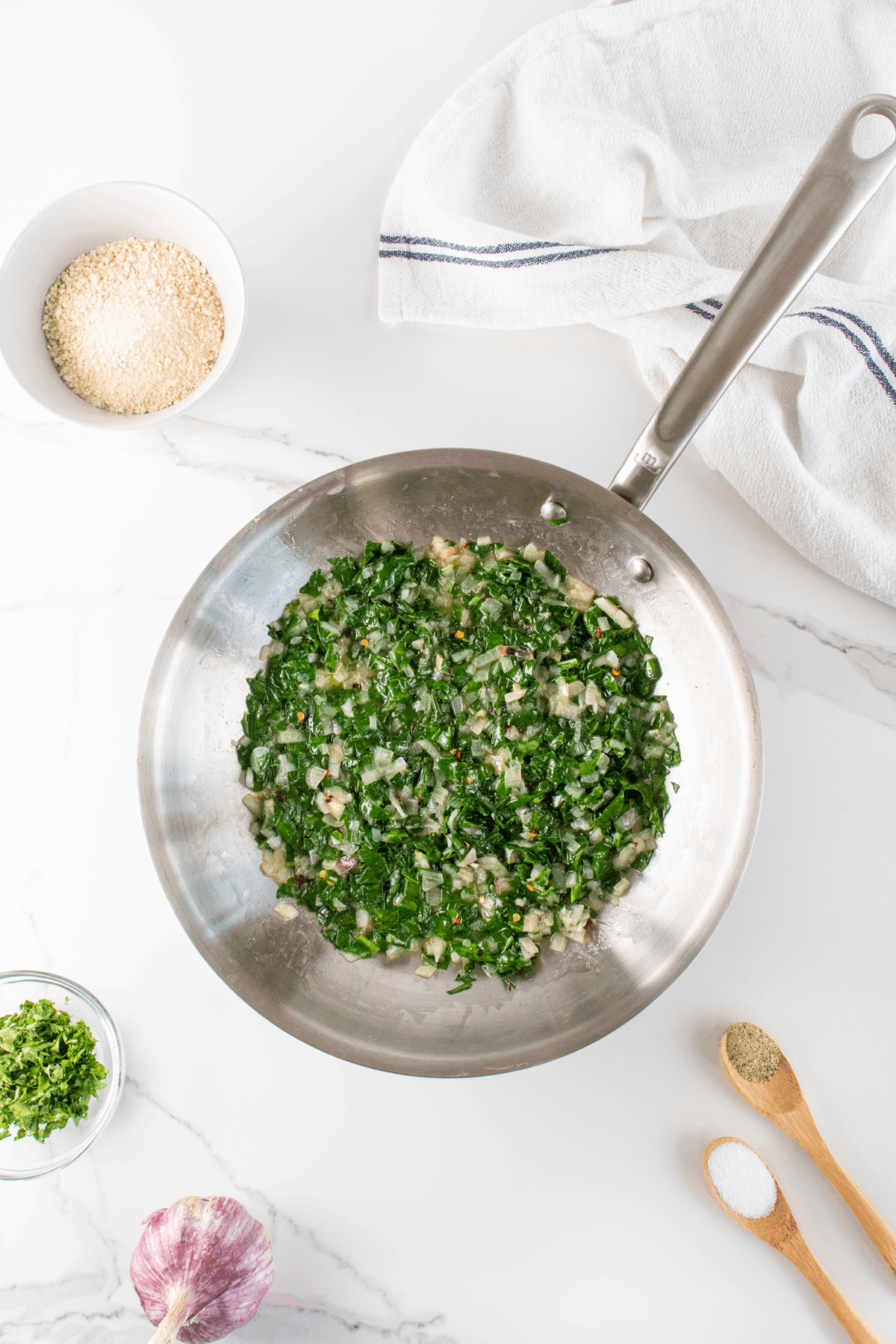A stainless steel pan with chopped greens, onions, and garlic cooking on a white marble surface, surrounded by a bowl of grains, a small bowl of chopped herbs, garlic, and wooden spoons with salt and pepper.