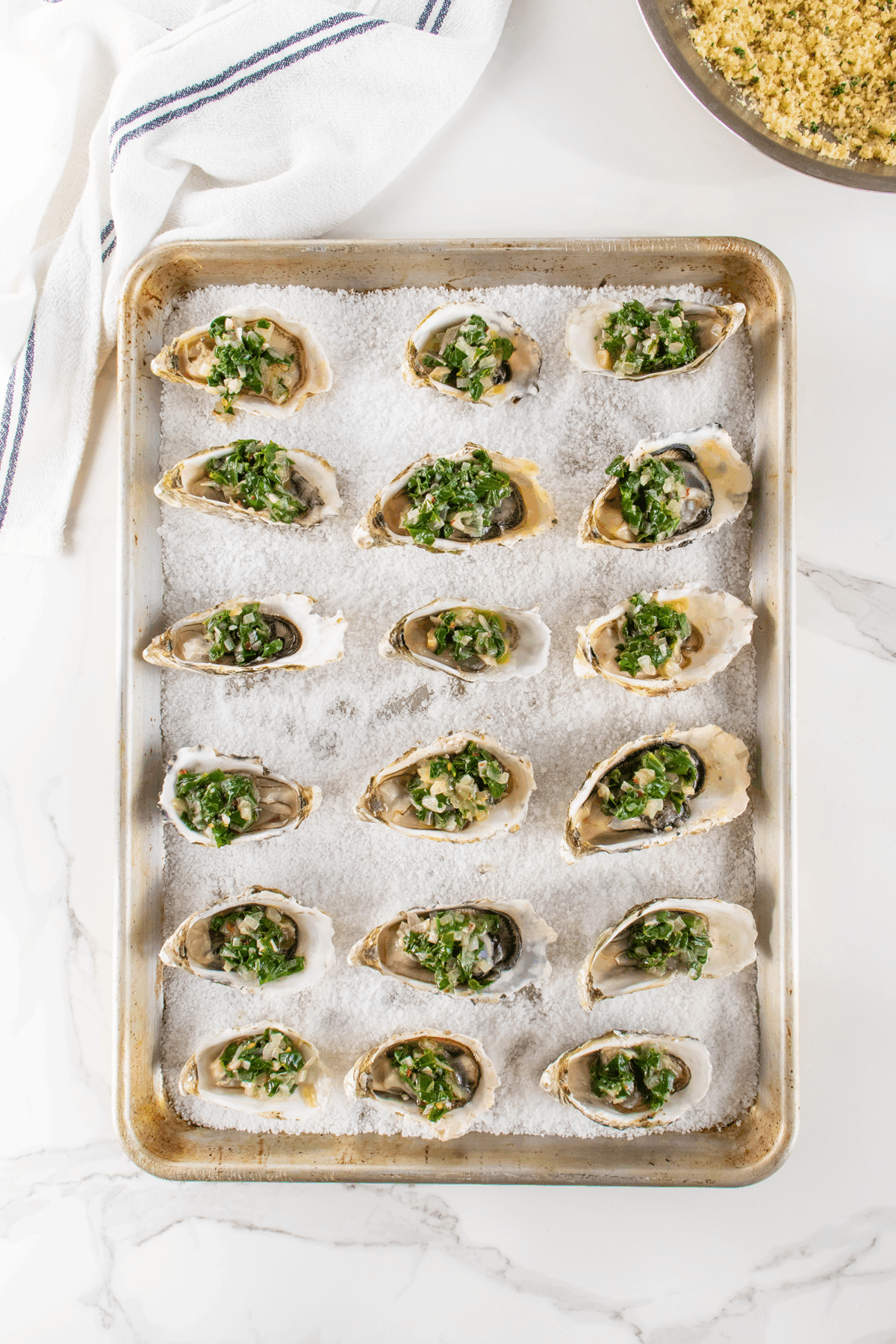 A baking sheet filled with oysters on the half shell, each topped with chopped green herbs, sits on a layer of coarse salt. A white kitchen towel and a bowl of breadcrumb mixture are nearby on a marble countertop.