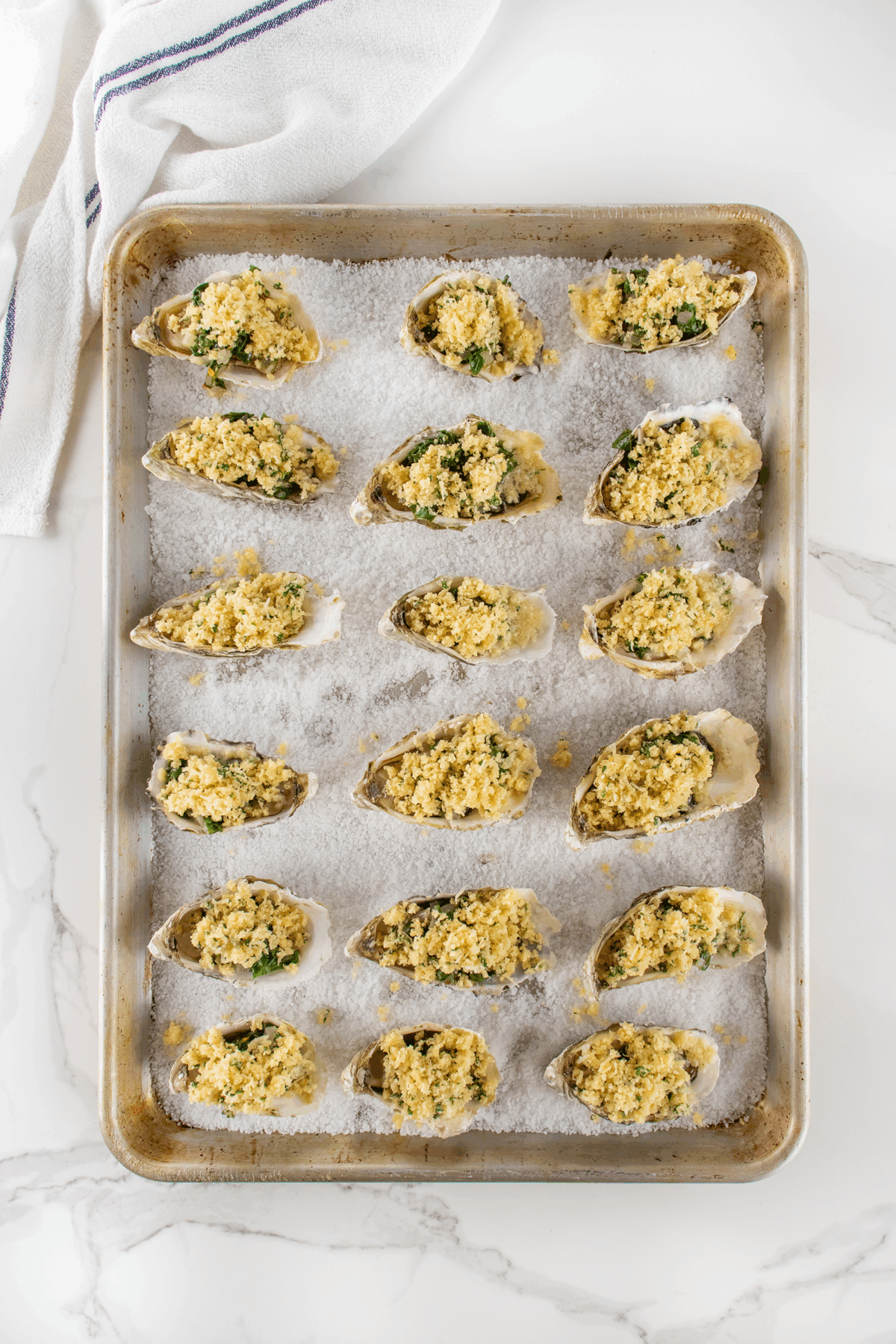 A baking sheet holds fifteen oysters on the half shell, topped with a breadcrumb and herb mixture, arranged on a layer of coarse salt. A white kitchen towel with blue stripes is in the upper left corner.