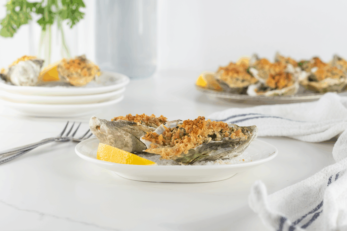 A close-up of baked oysters with a golden breadcrumb topping, served on a white plate with a lemon wedge. More oysters are visible in the background along with a white napkin and forks.