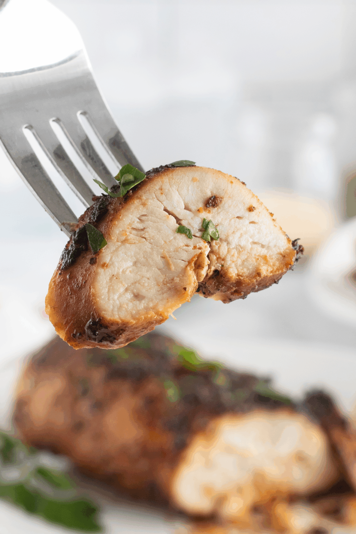 A close-up of a fork holding a piece of juicy, cooked chicken breast garnished with chopped herbs, with more chicken and parsley visible blurred in the background.