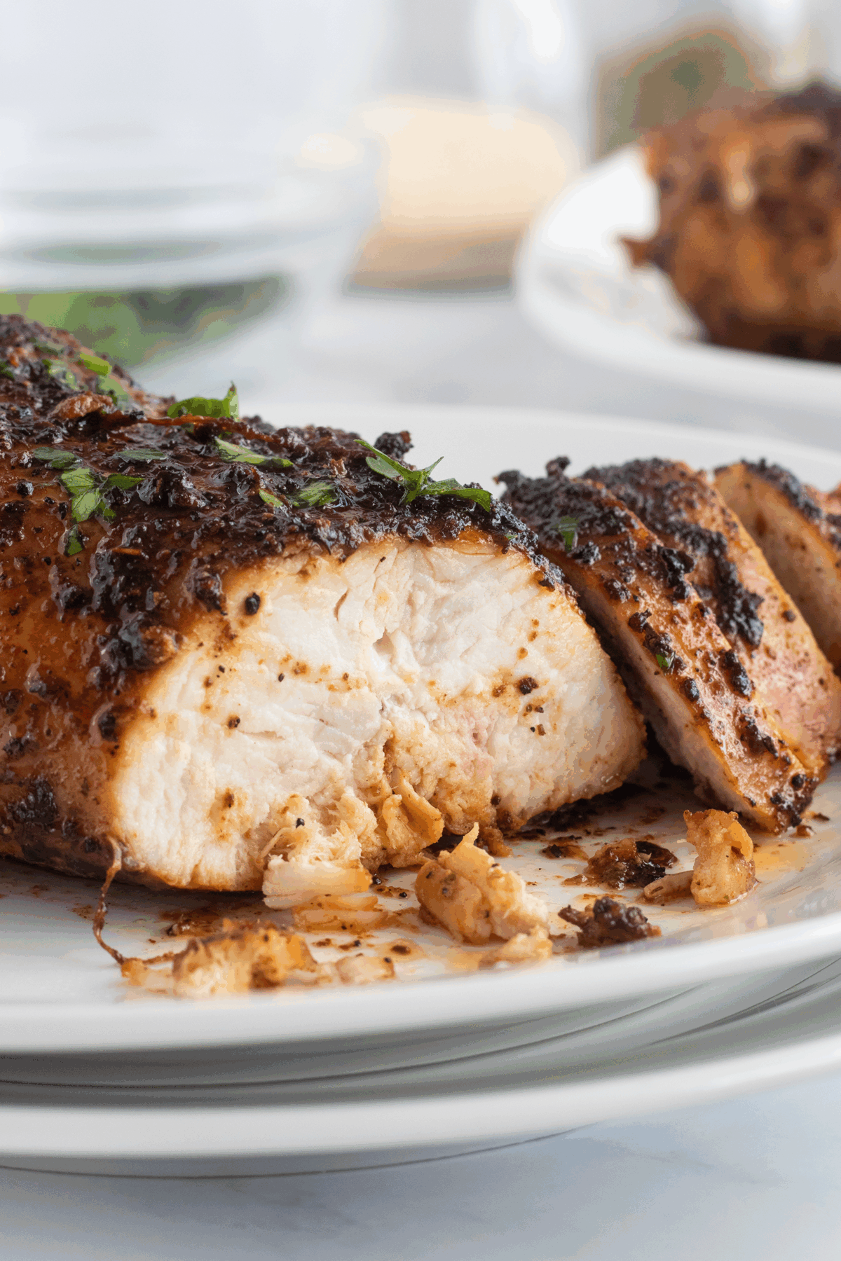 Close-up of sliced, juicy, seasoned chicken breast on a white plate, garnished with herbs. The chicken appears tender and well-cooked, with flavorful spices and juices visible.