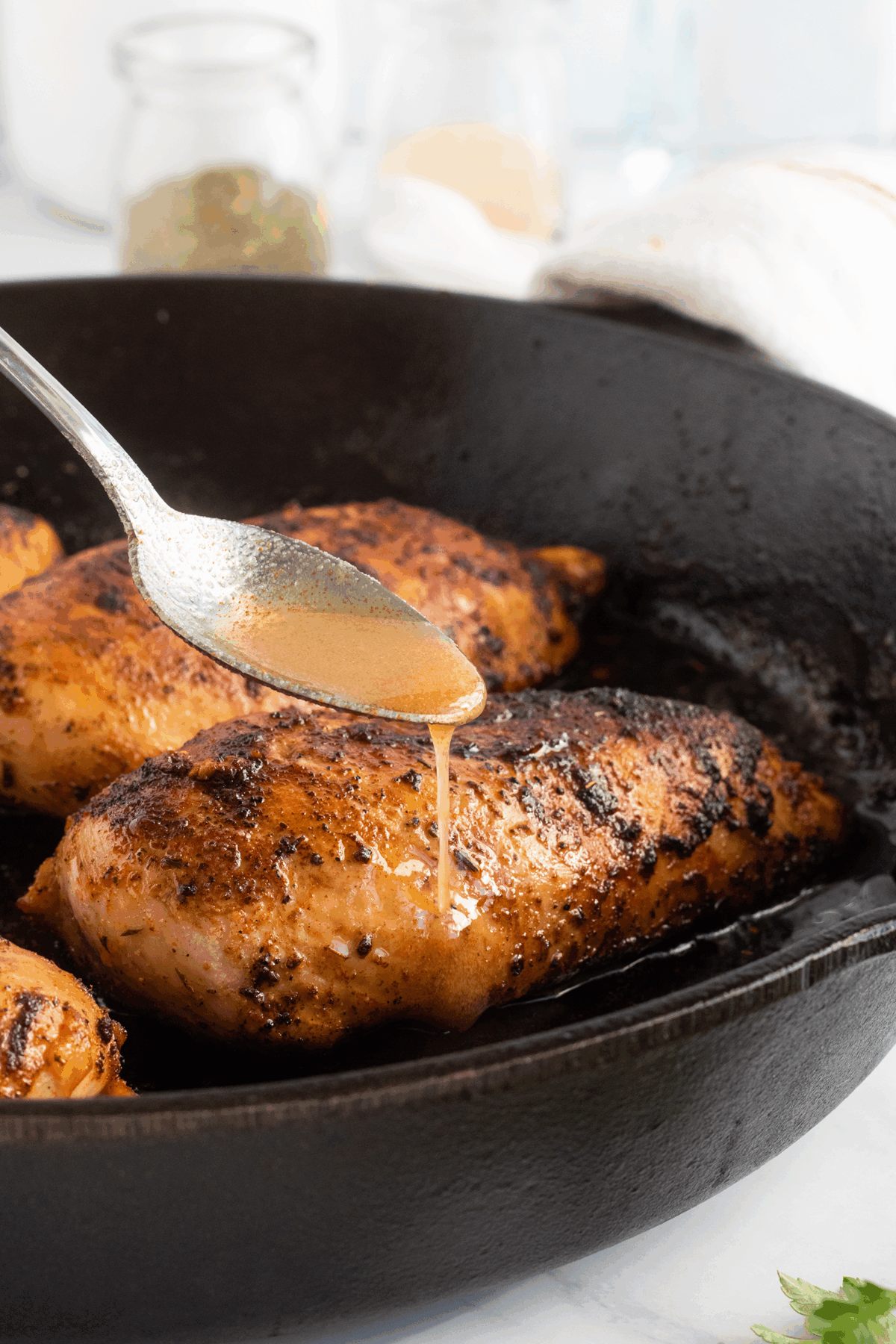 A spoon drizzles sauce over a seasoned, cooked chicken breast in a black cast iron skillet. Other breasts are visible, and the background shows jars and a towel out of focus.