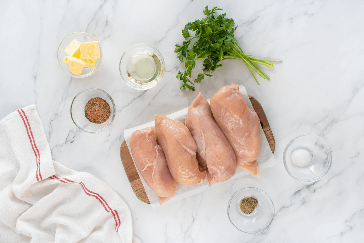 Four raw chicken breasts on a cutting board, surrounded by small bowls of butter, oil, spices, salt, and pepper, with a bunch of parsley and a white kitchen towel on a marble countertop.