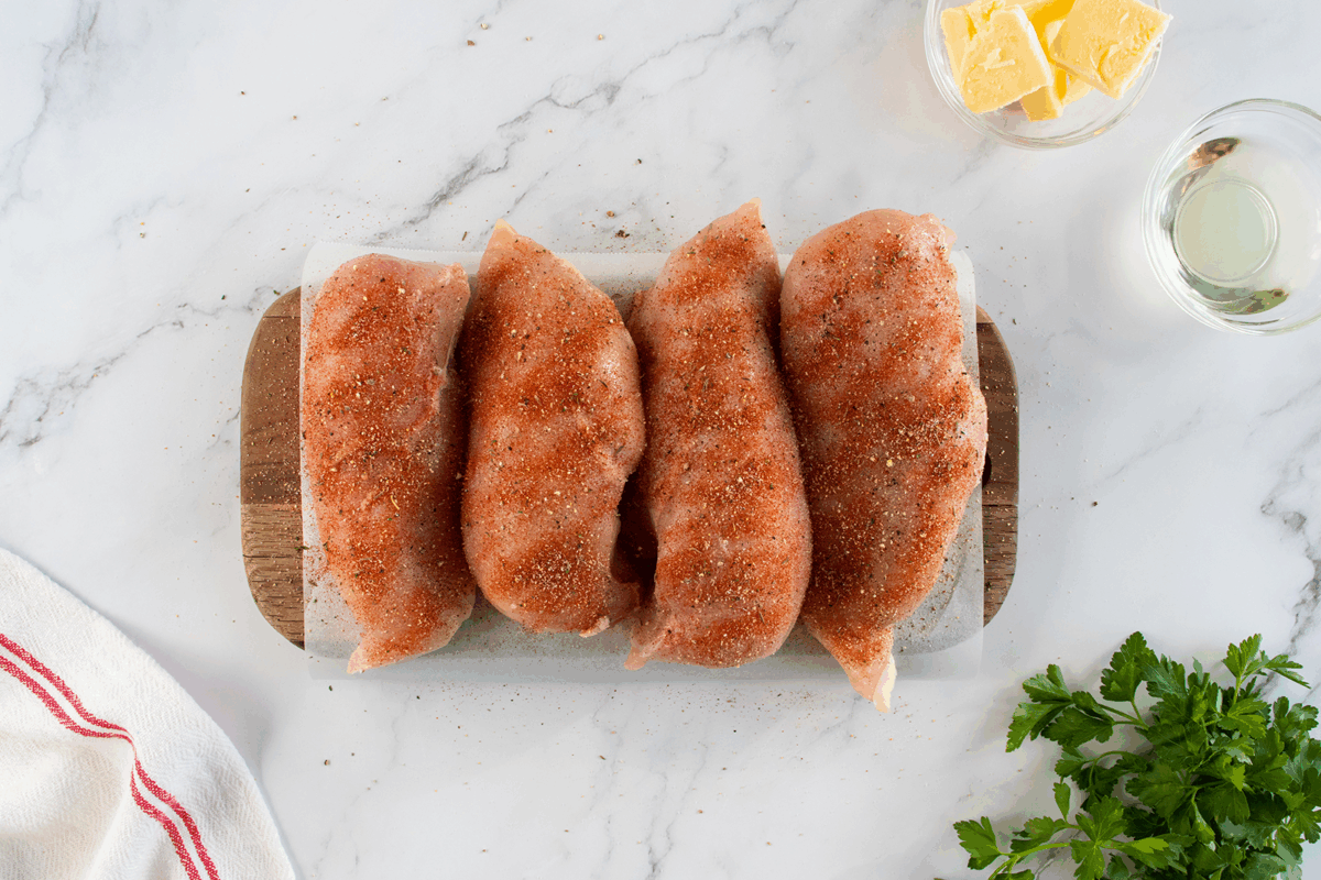 Four raw, seasoned chicken breasts are arranged in a row on a small cutting board. Nearby are a bowl of butter cubes, a glass of liquid, a towel, and some fresh parsley on a white marble surface.
