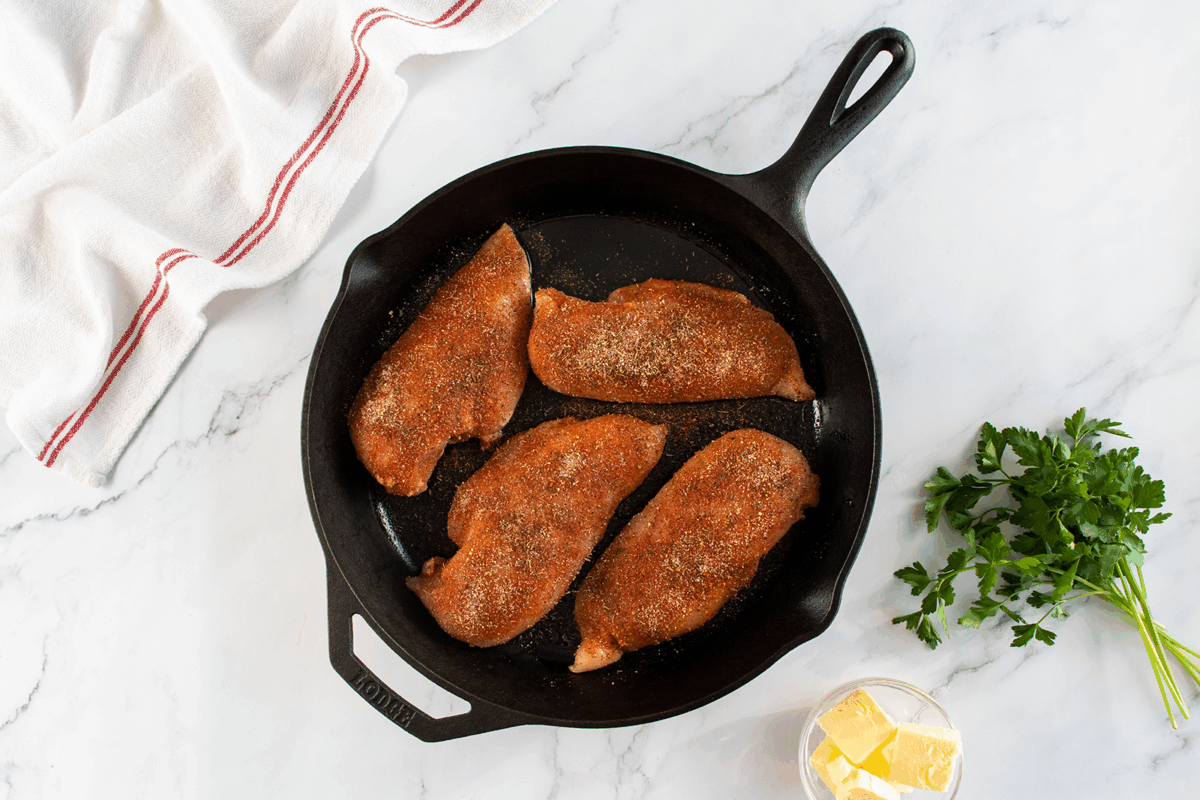 A black cast iron skillet with four seasoned raw chicken breasts on a marble surface, next to a bunch of fresh parsley, a small bowl of butter, and a white towel with red stripes.