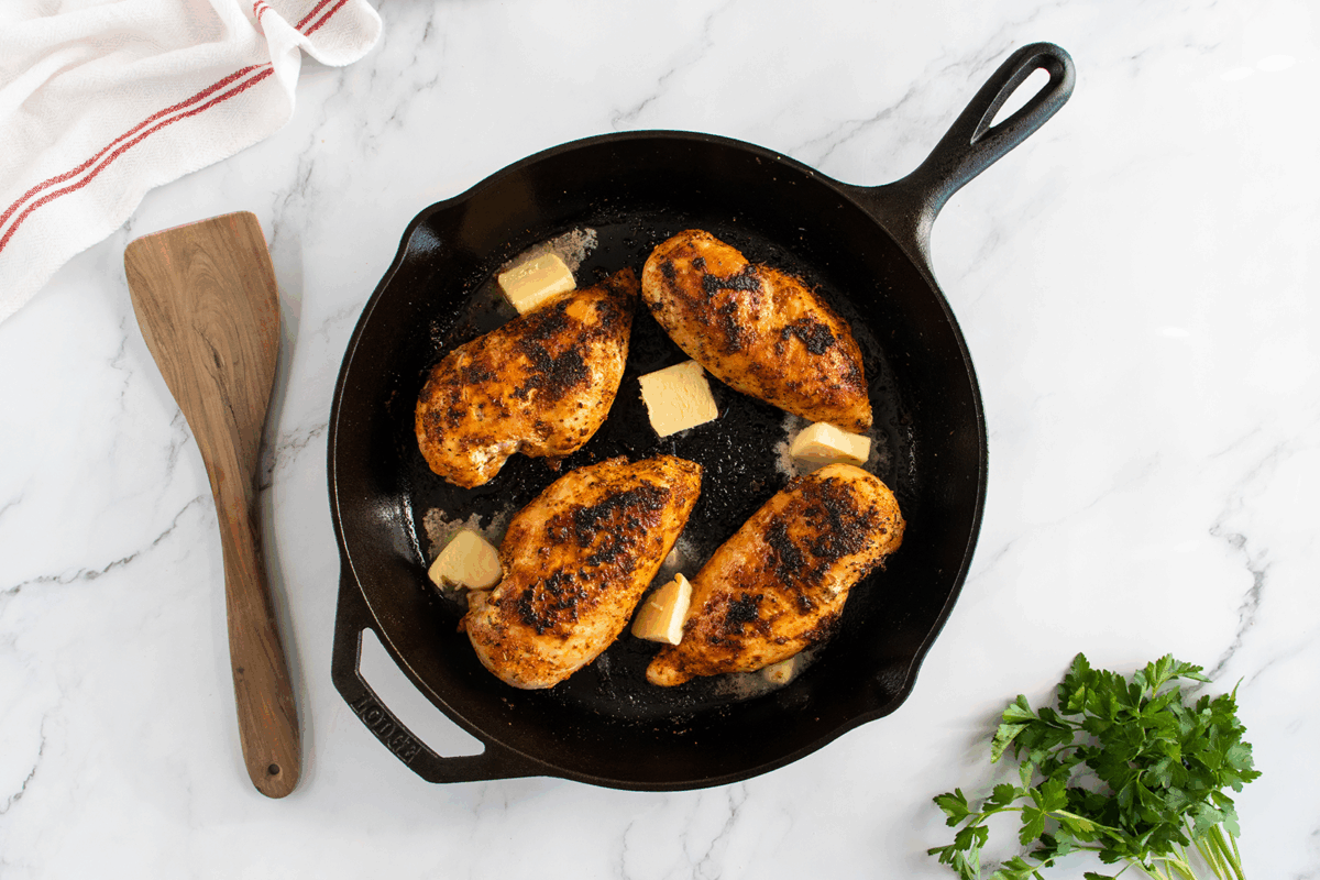 A cast iron skillet with four seasoned, cooked chicken breasts and melting butter sits on a white marble surface. A wooden spatula and a bunch of fresh parsley are nearby.