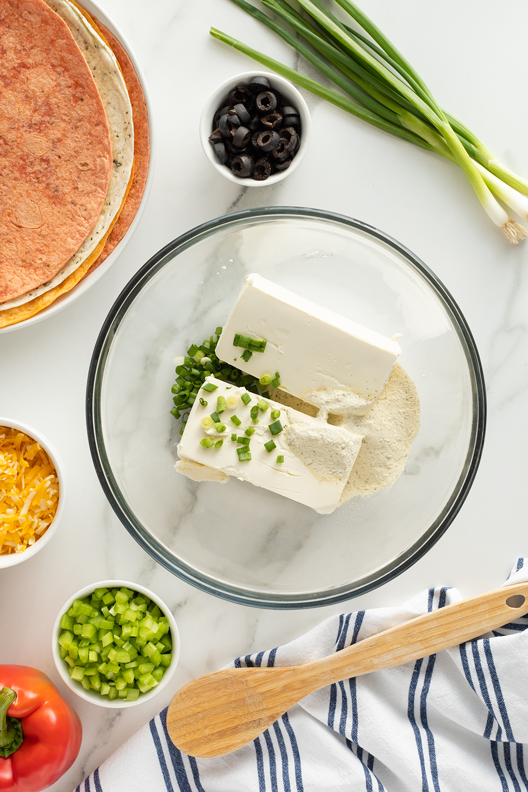 A glass bowl with cream cheese, chopped green onions, and seasoning sits on a white surface surrounded by chopped celery, shredded cheese, sliced olives, tortillas, a tomato, green onions, and a wooden spoon on a striped towel.