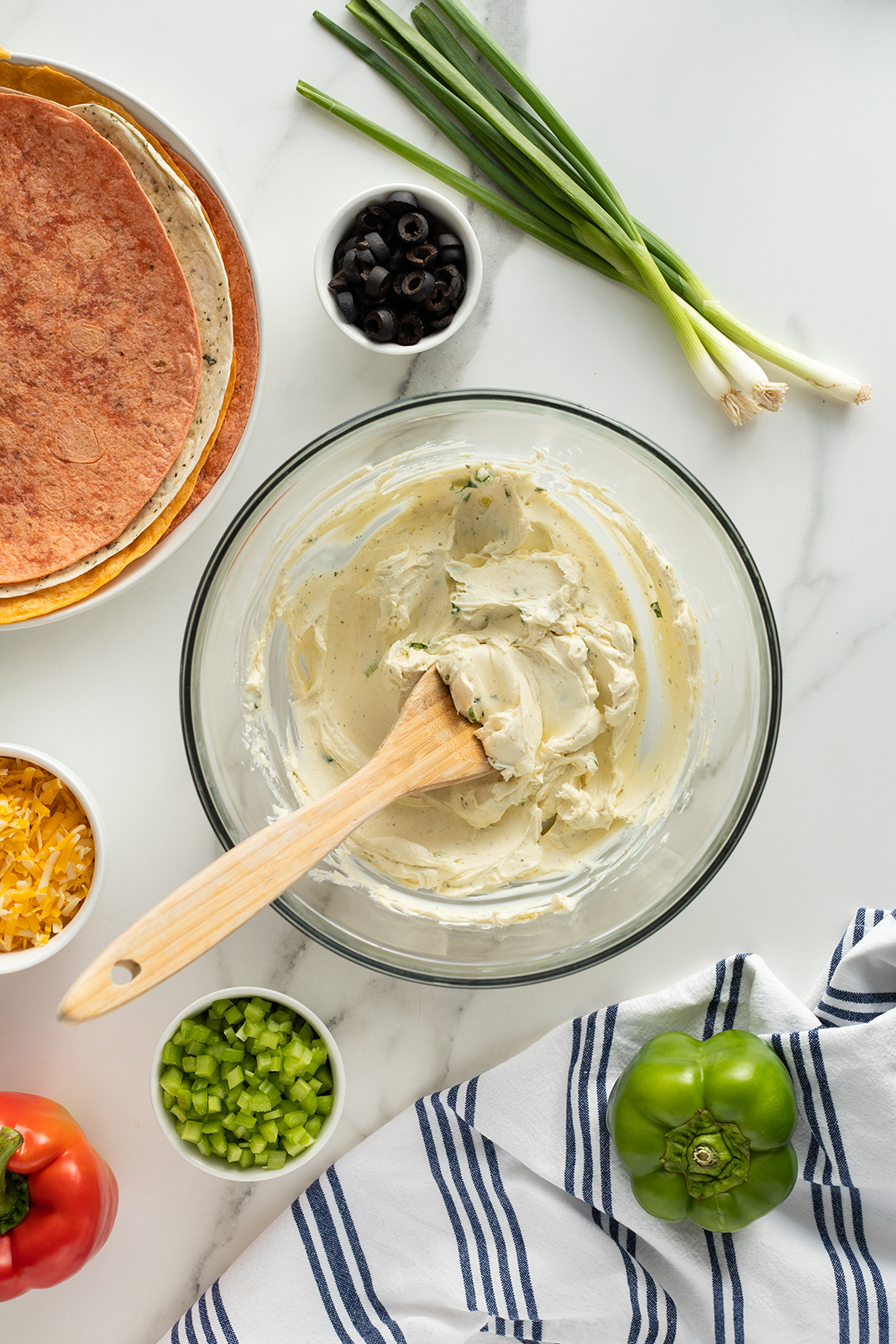 A glass bowl of creamy mixture with a wooden spatula is surrounded by chopped green peppers, shredded cheese, sliced olives, green onions, bell pepper, tortillas, and a striped kitchen towel on a white surface.