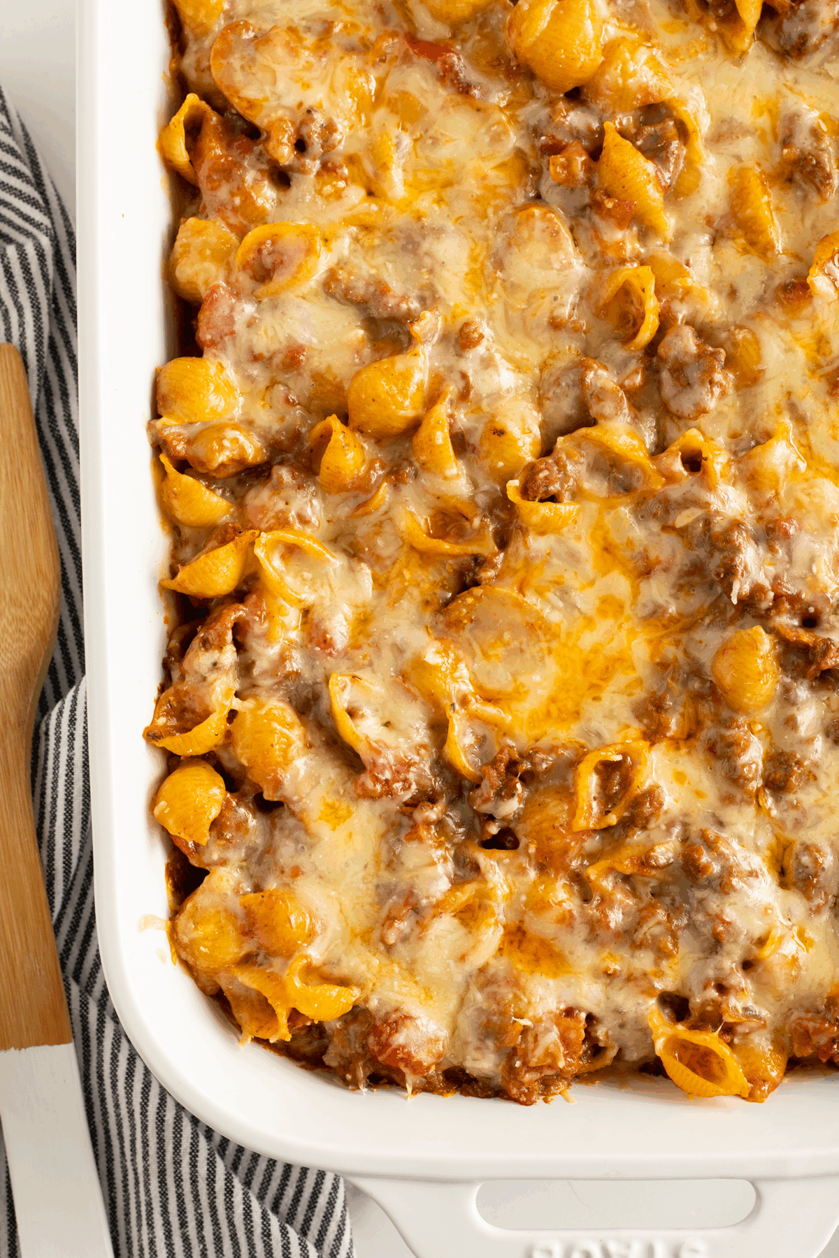 A close-up of a baked pasta casserole with shell pasta, ground meat, tomato sauce, and melted cheese in a white baking dish, next to a striped kitchen towel and wooden cutting board.