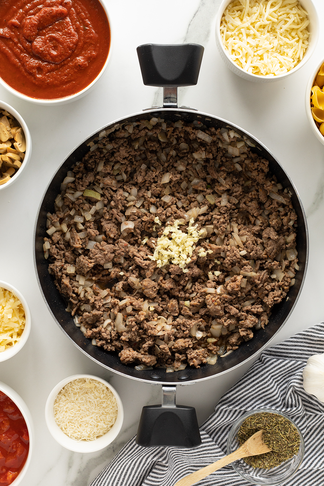 Overhead view of a skillet filled with cooked ground beef and chopped onions, topped with minced garlic. Surrounding the skillet are bowls of tomato sauce, shredded cheese, uncooked rice, seasonings, and a striped towel.