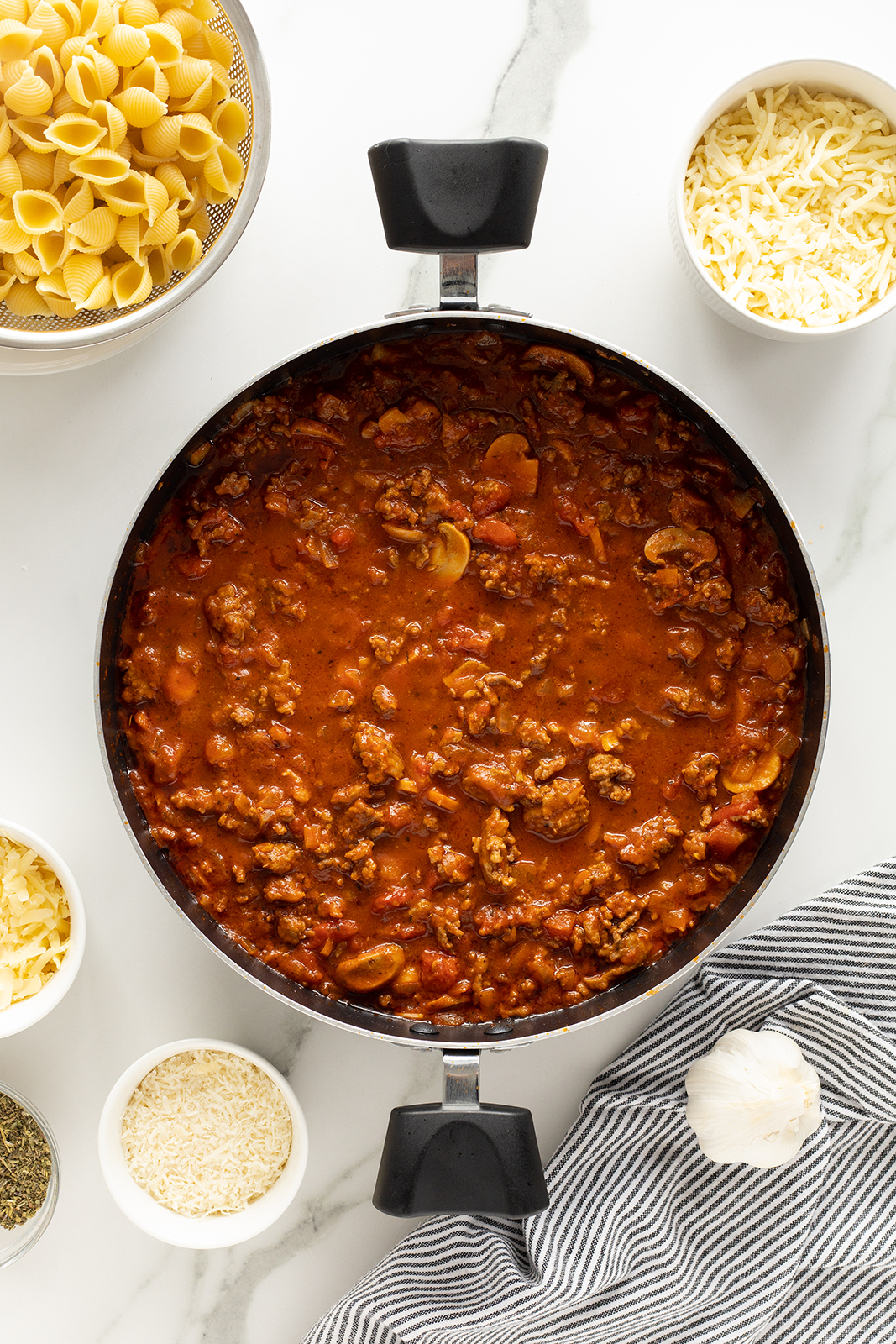 A pot of meat and tomato sauce with mushrooms on a white surface, surrounded by bowls of dry pasta shells, shredded cheese, white rice, sliced garlic, and dried herbs; a striped cloth sits nearby.