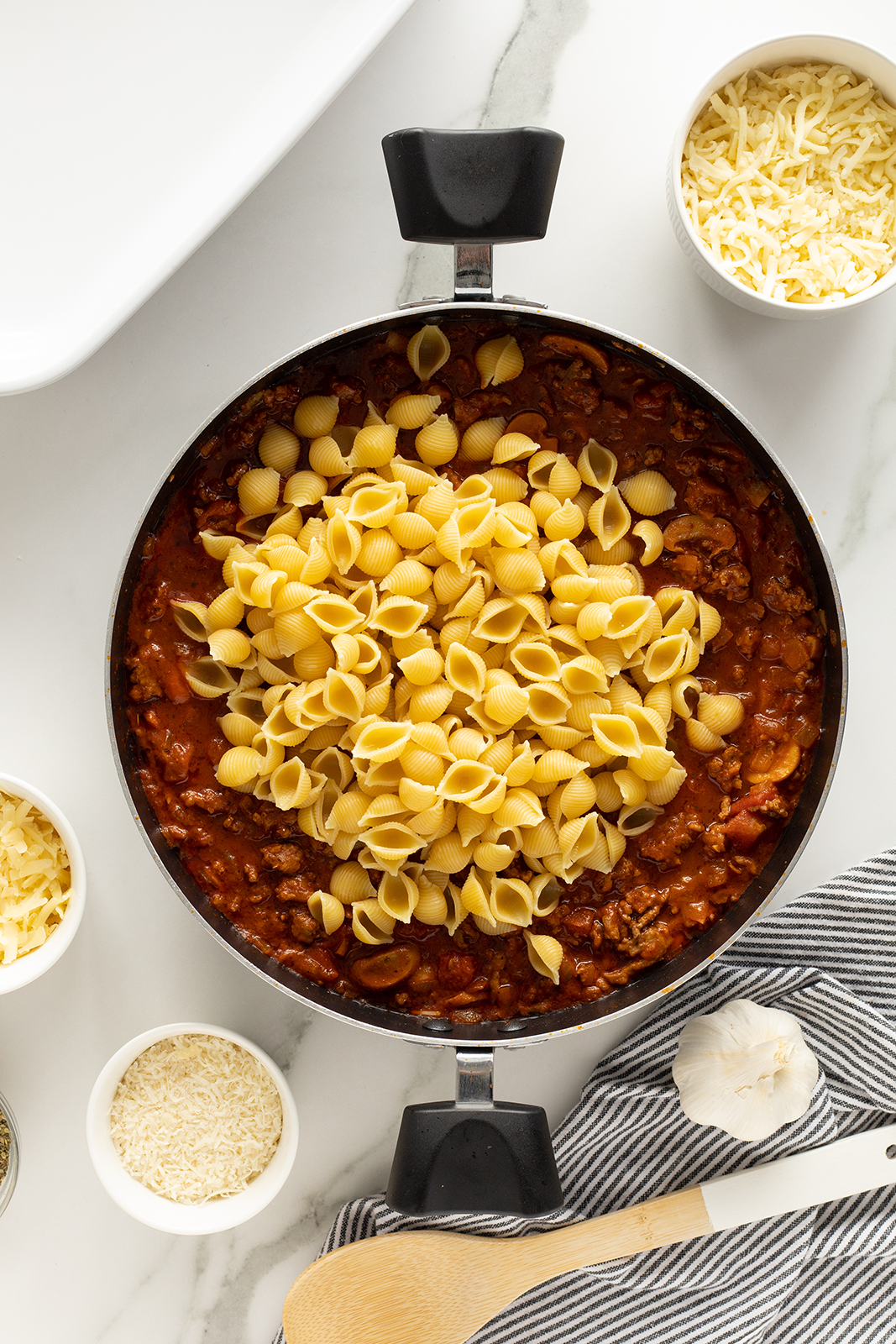 A skillet filled with tomato meat sauce is topped with uncooked shell pasta. Surrounding the skillet are bowls of shredded cheese, uncooked rice, a head of garlic, and a wooden spoon on a striped towel.