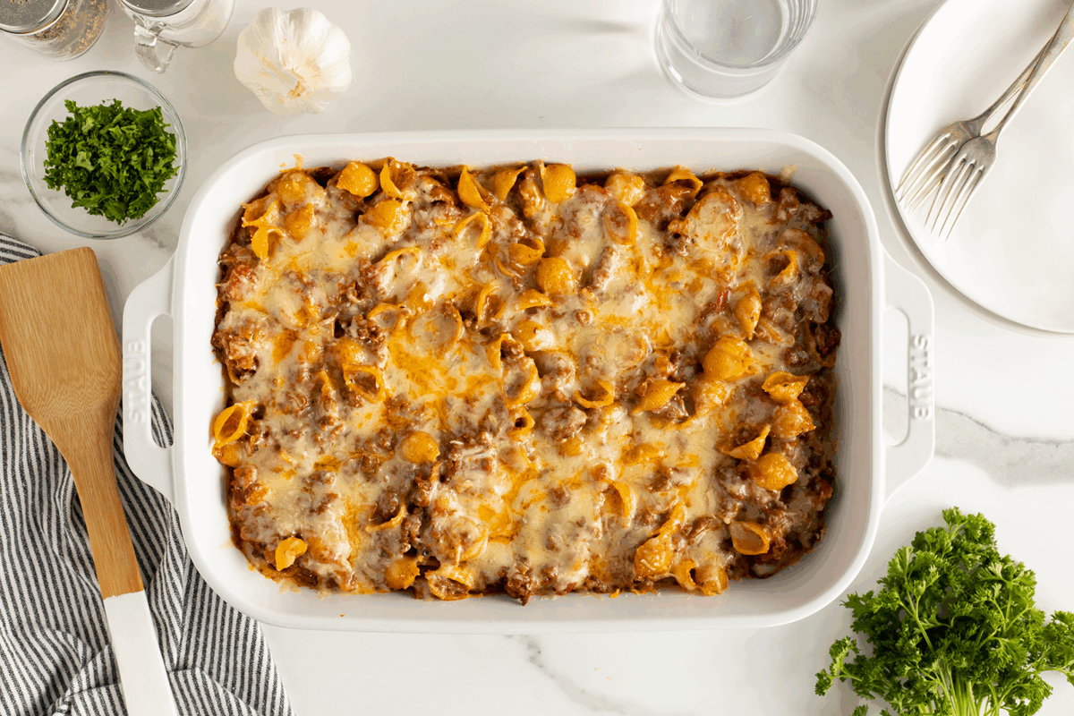 A white baking dish filled with cheesy baked pasta shells in a meat sauce sits on a marble countertop, surrounded by fresh parsley, a wooden spatula, a fork and plate, garlic, salt and pepper, and a glass of water.