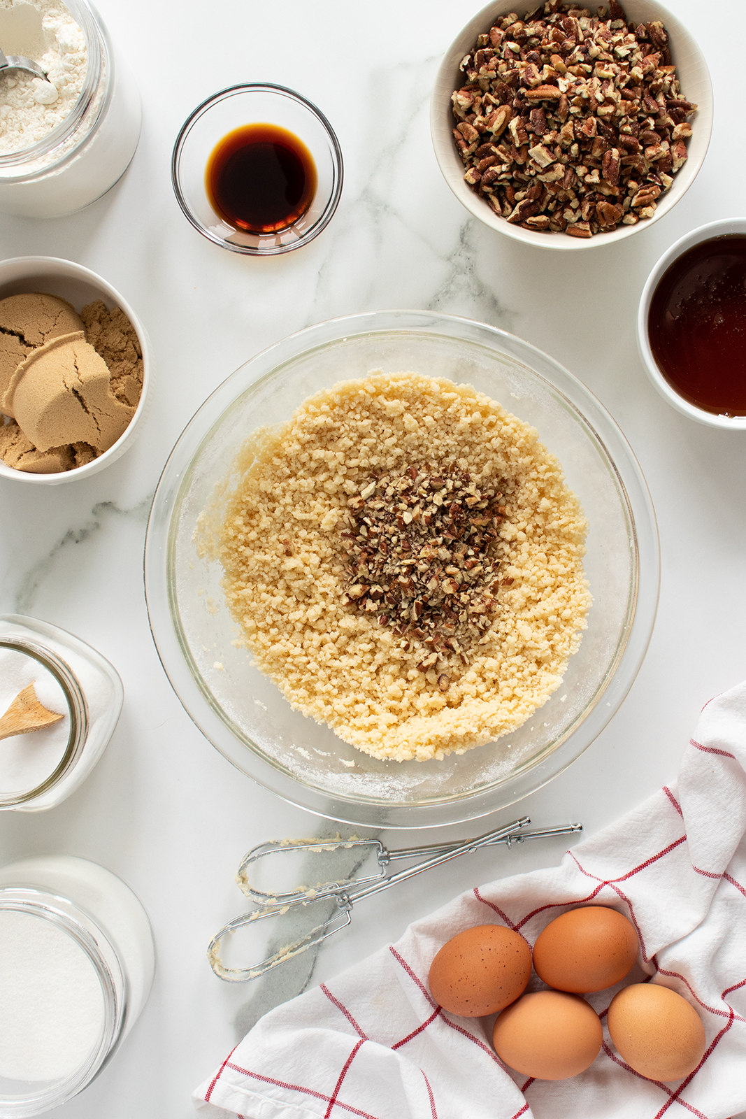 A bowl of cookie dough topped with chopped pecans is surrounded by eggs, a whisk, sugar, flour, brown sugar, vanilla, syrup, and a red-and-white towel on a white countertop.