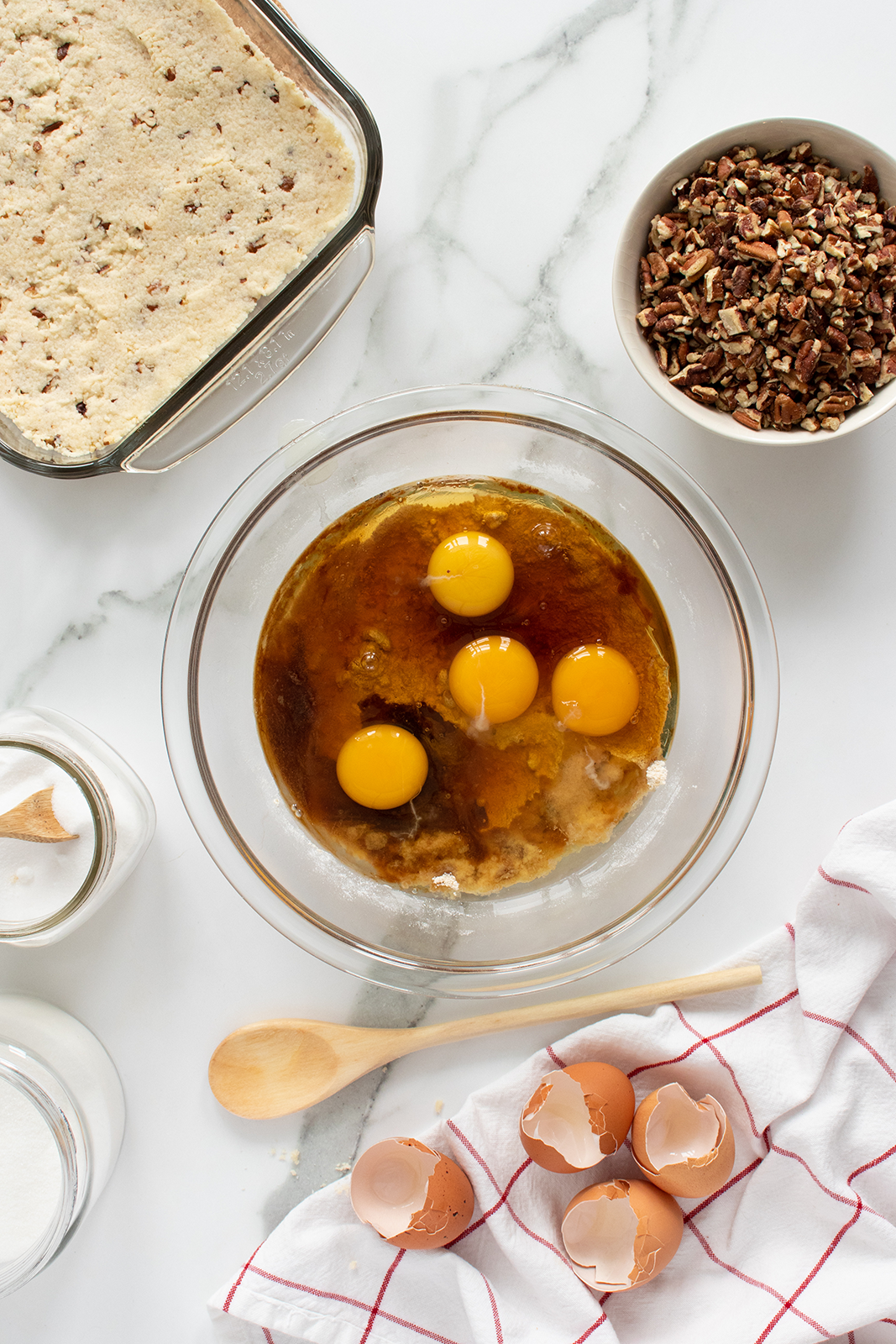 A glass bowl containing four eggs and vanilla sits on a marble countertop surrounded by a baking dish with crust, chopped pecans, egg shells, a wooden spoon, white kitchen towel, and jars of sugar and flour.
