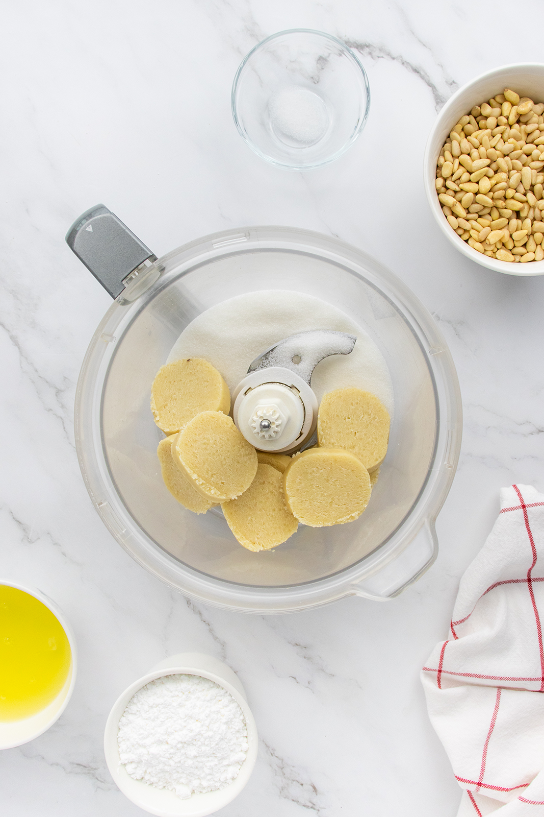 A food processor with six slices of dough inside sits on a marble counter, surrounded by bowls of olive oil, powdered sugar, a small bowl of salt, pine nuts, and a red-striped kitchen towel.