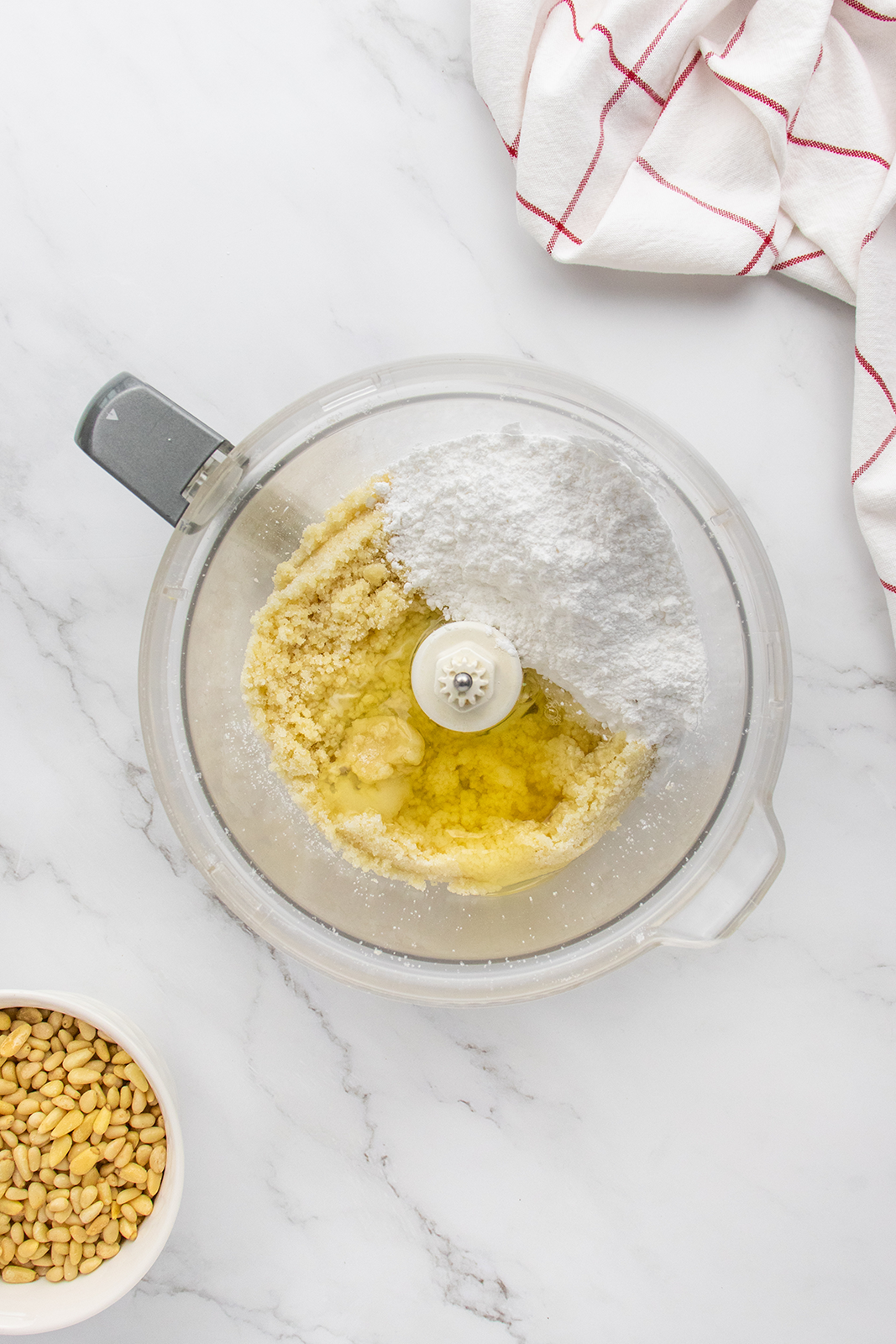 A food processor bowl with butter, sugar, and flour inside sits on a marble countertop, next to a white bowl of pine nuts and a white towel with red stripes.