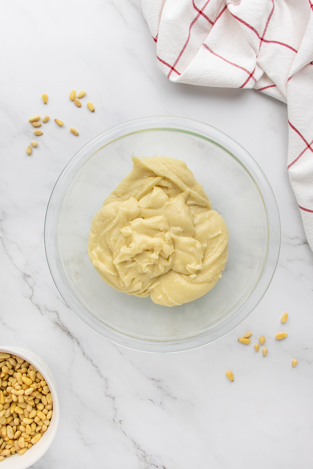 A glass bowl filled with creamy, pale hummus sits on a white marble surface. Scattered pine nuts and a red-striped white kitchen towel are nearby, along with a small bowl of more pine nuts.