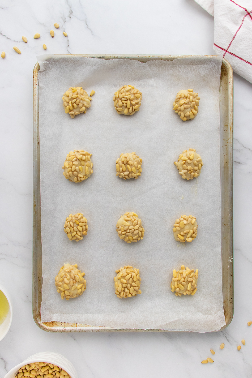 A baking sheet lined with parchment paper holds twelve unbaked, round cookies coated with pine nuts, evenly spaced in rows. Nearby, a bowl of pine nuts and a white towel with a red stripe are visible.