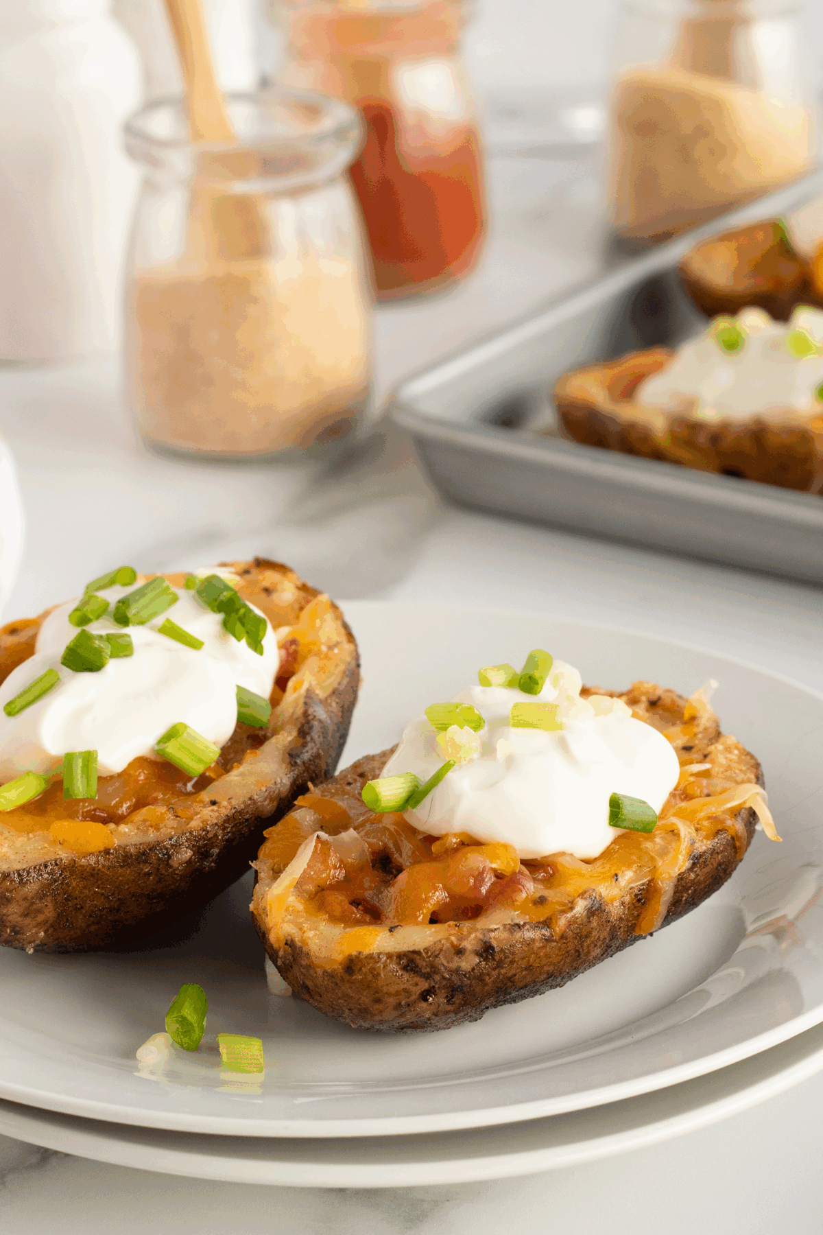 Two loaded potato skins topped with melted cheese, sour cream, and chopped green onions on a white plate. A baking tray with more potato skins and jars of spices are in the background.