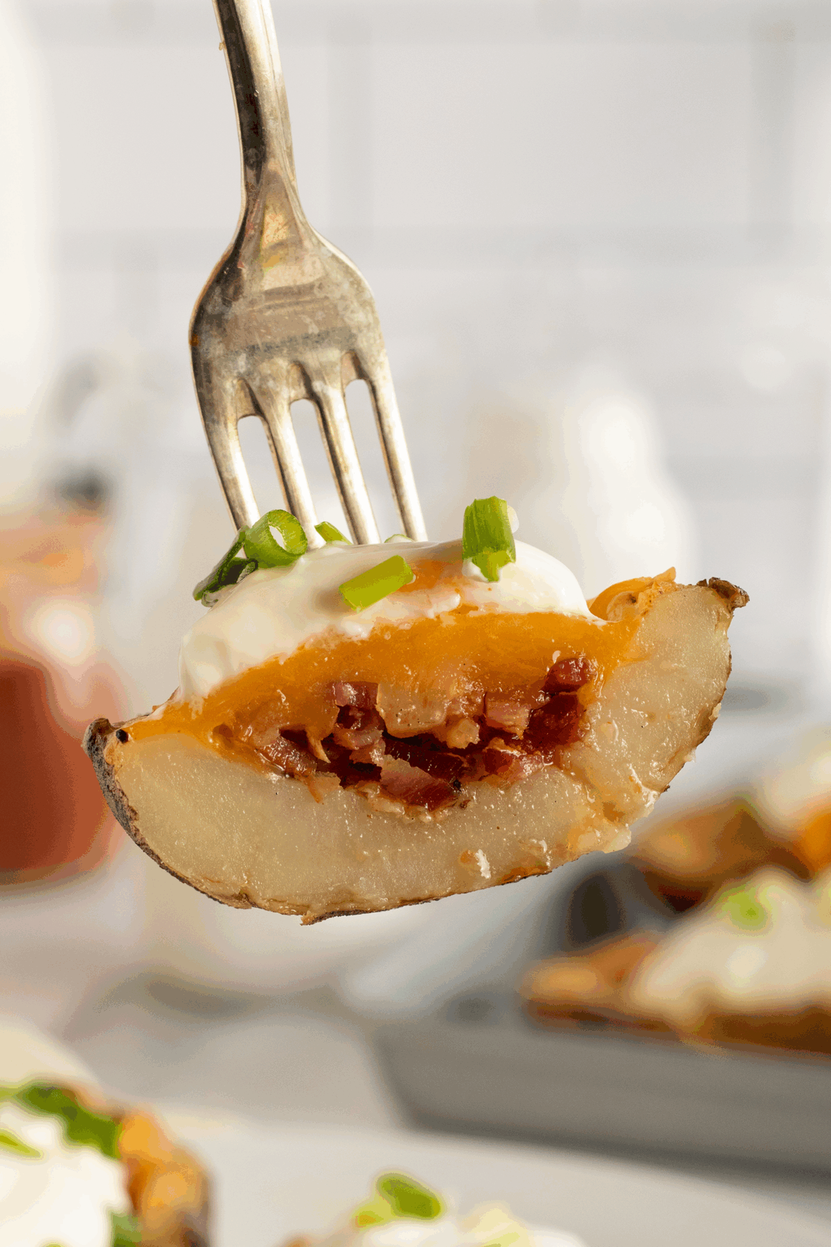 A close-up of a fork holding a potato skin filled with melted cheese, bacon bits, sour cream, and chopped green onions. More potato skins are visible on a tray in the background.