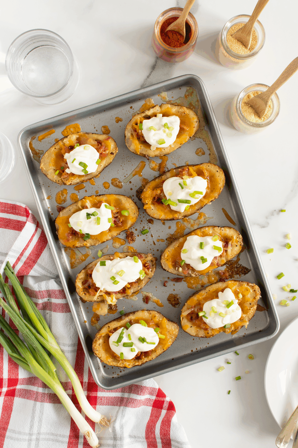 A baking sheet with loaded potato skins topped with melted cheese, sour cream, and chopped green onions sits on a white surface, next to a red-striped towel, green onions, and jars of spices.
