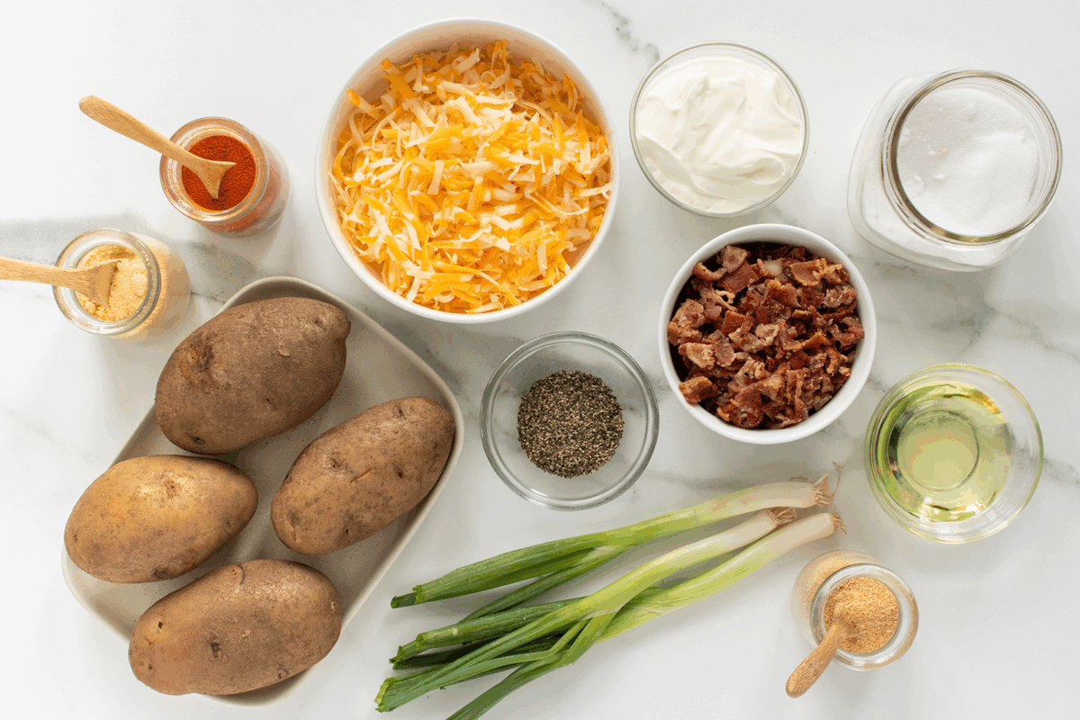 Ingredients for loaded baked potatoes arranged on a white surface: four russet potatoes, shredded cheese, cooked bacon bits, green onions, black pepper, sour cream, oil, salt, paprika, and garlic powder.