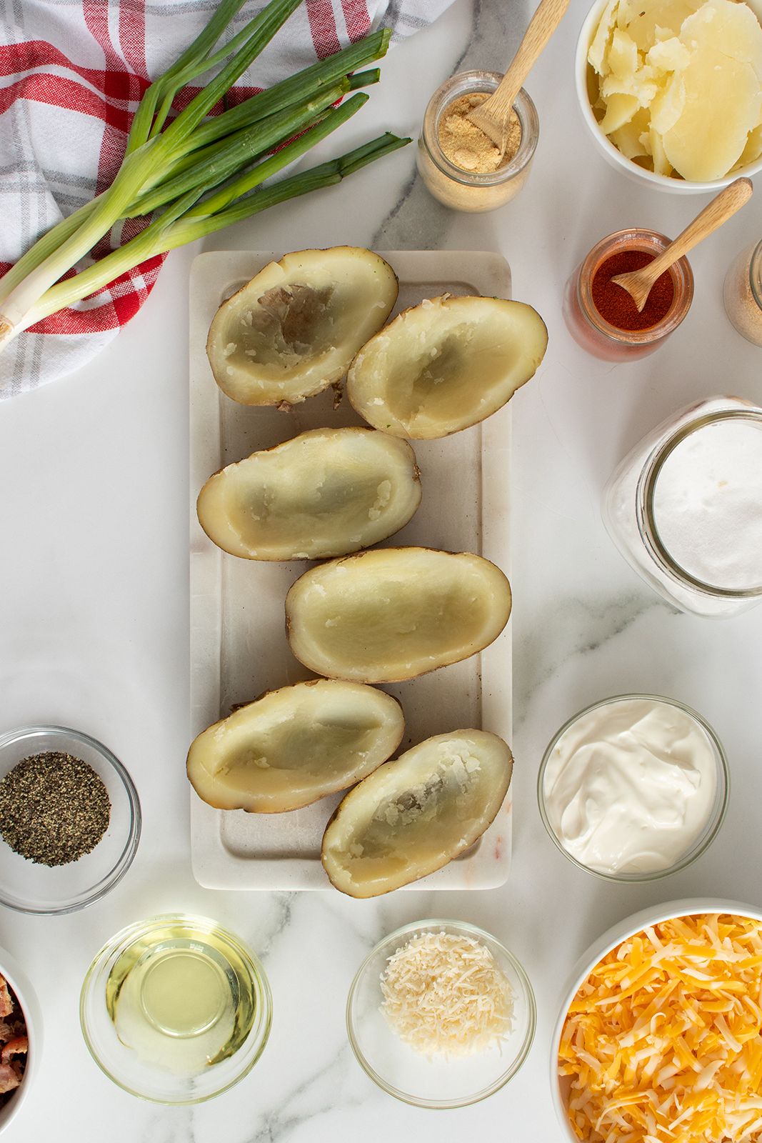 A marble countertop with halved baked potatoes on a cutting board, surrounded by bowls of shredded cheese, sour cream, onion, spices, oil, and green onions, ready for making stuffed potatoes.