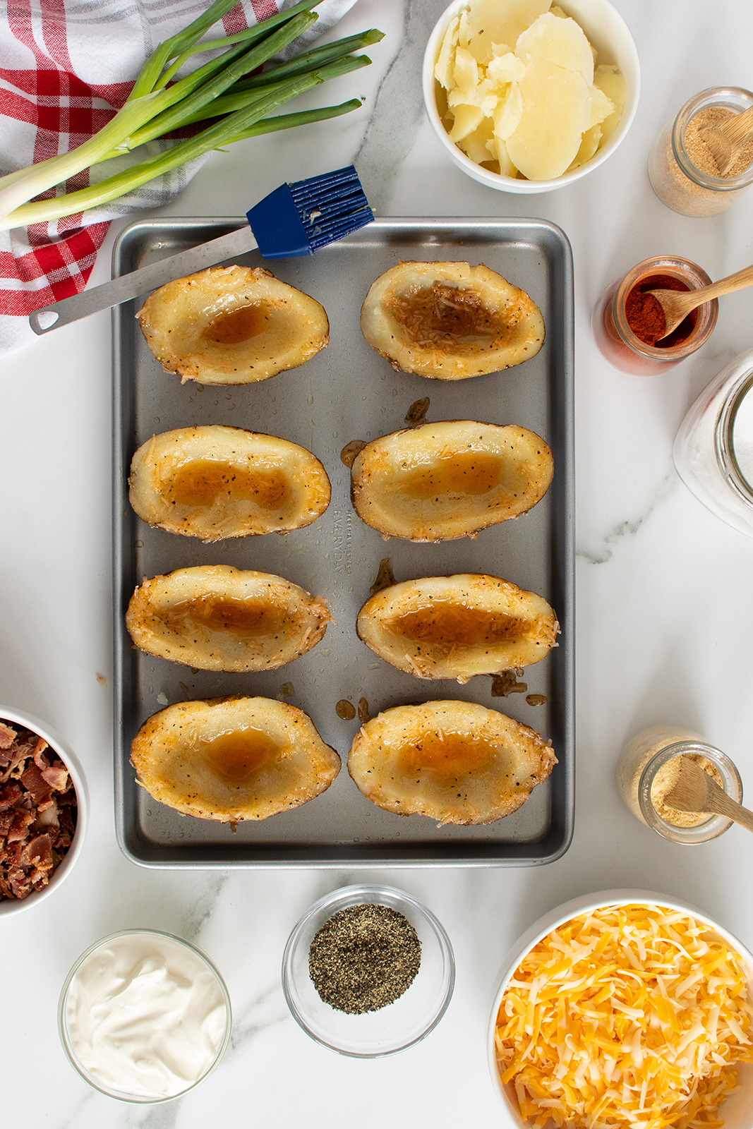 A baking tray with eight hollowed potato skins sits on a marble counter, surrounded by bowls of shredded cheese, bacon bits, sour cream, green onions, sliced potatoes, spices, and a blue basting brush.