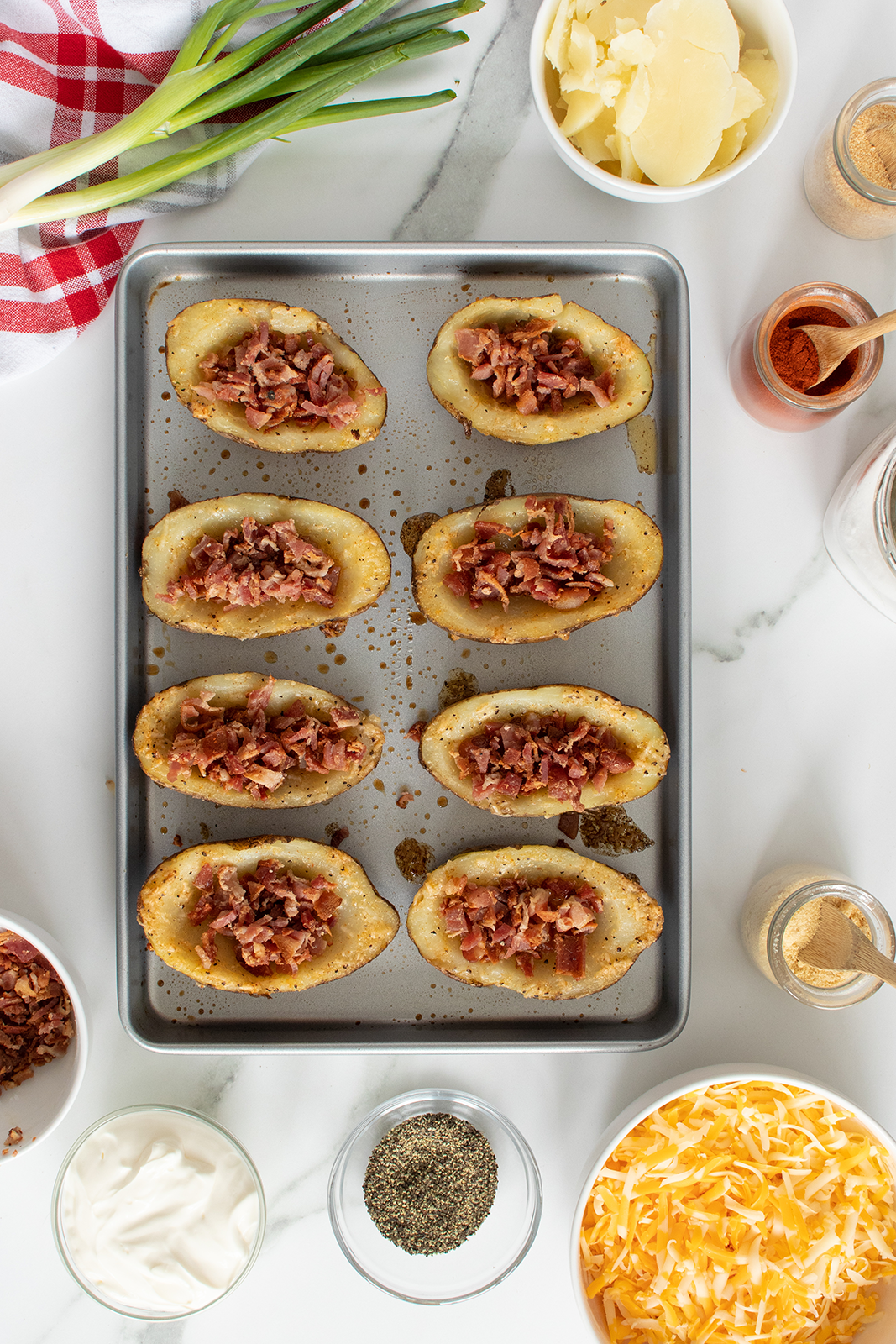 A baking tray holds eight potato skins filled with crispy bacon bits. Surrounding the tray are bowls of shredded cheese, black pepper, sour cream, spices, green onions, and a red-checkered napkin.
