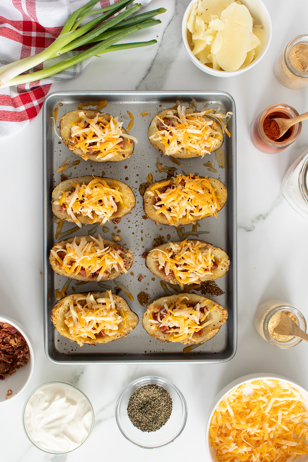 A baking tray with eight potato halves topped with shredded cheese, surrounded by bowls of cheese, sour cream, bacon bits, sliced green onions, pepper, and seasonings on a marble countertop.