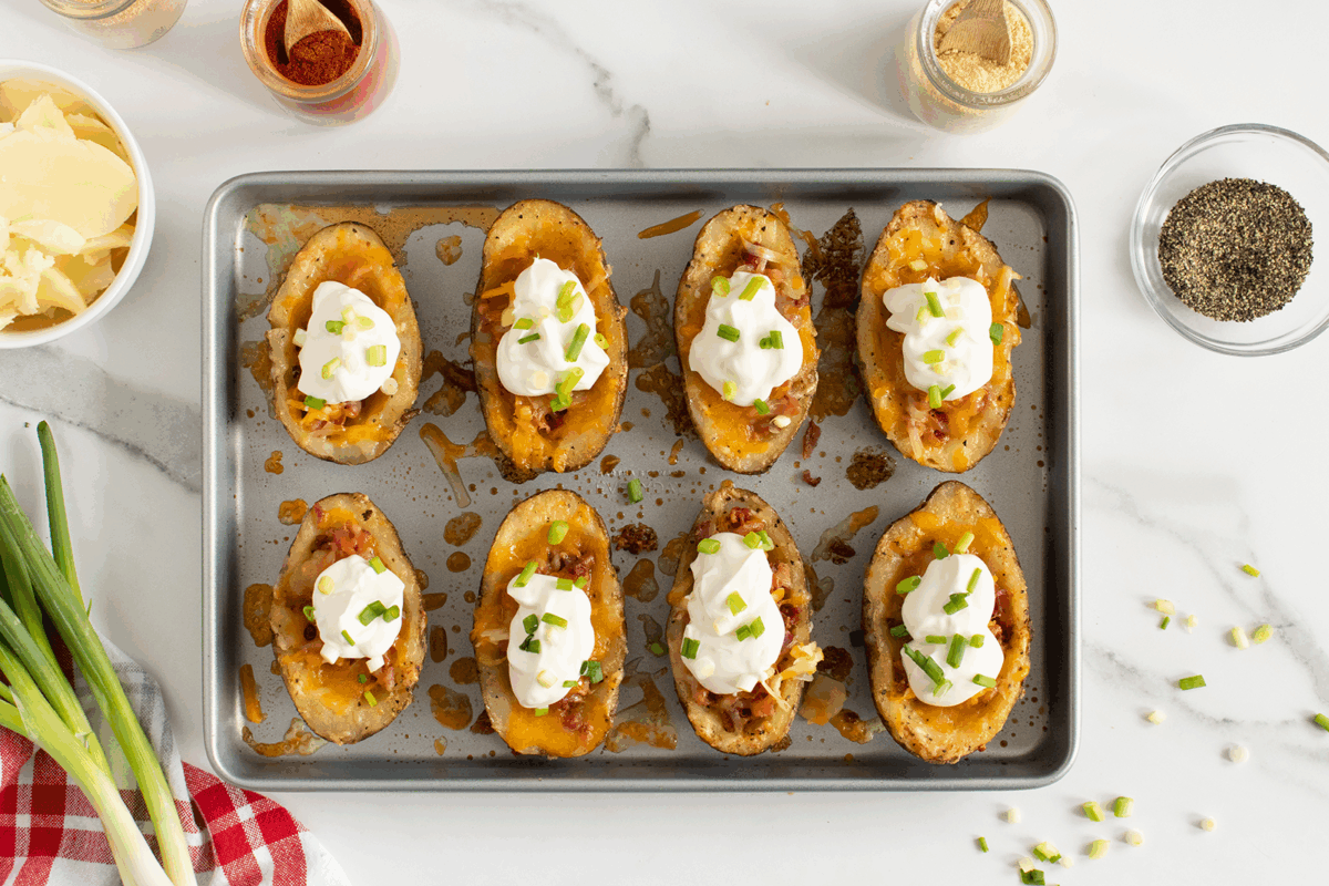 A baking sheet with eight loaded potato skins topped with melted cheese, bacon bits, sour cream, and chopped green onions. Seasonings and toppings are arranged around the tray on a white marble surface.