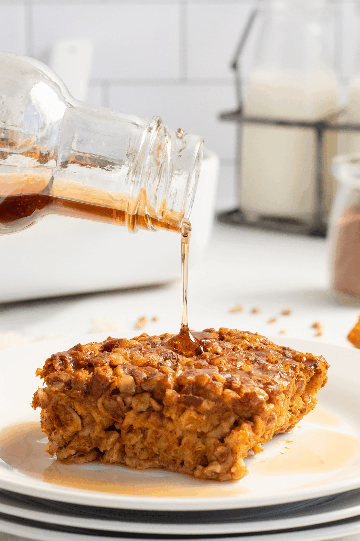 A close-up of syrup being poured from a glass bottle onto a piece of baked French toast casserole with chopped nuts, served on a white plate. Milk and kitchen items are blurred in the background.