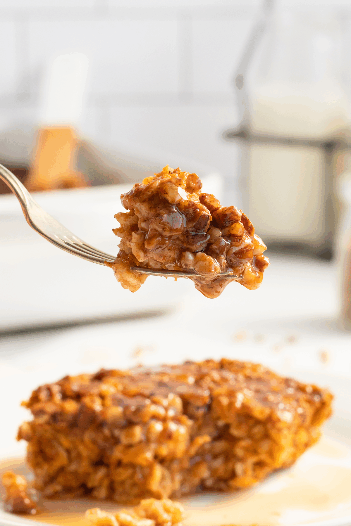 A close-up of a fork holding a bite of baked oatmeal, with a square of oatmeal and syrup on a white plate in the background. A glass of milk and a white dish are blurred in the background.