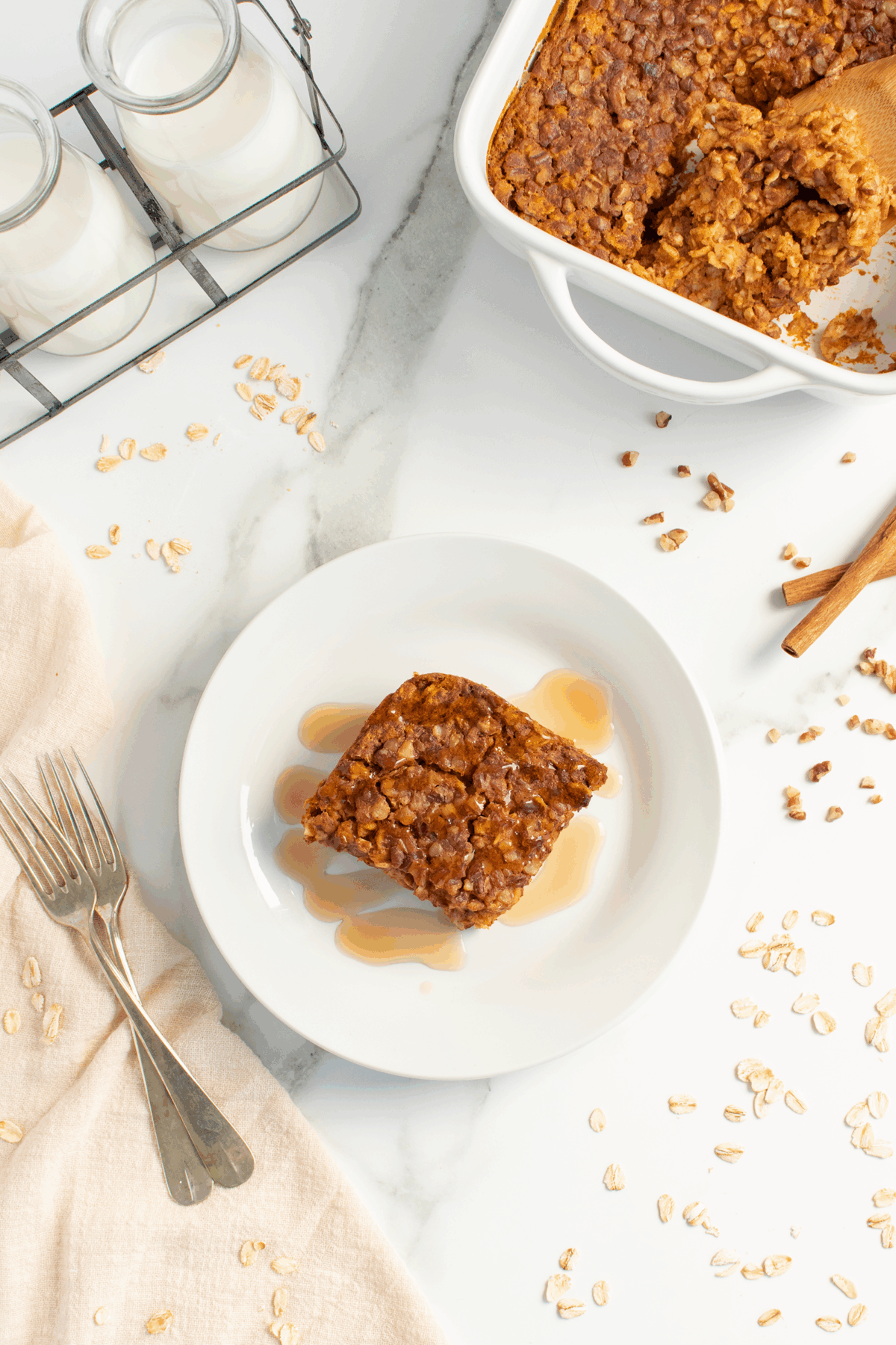 A square serving of baked oatmeal drizzled with syrup sits on a white plate. Nearby are forks, a beige napkin, a baking dish with more oatmeal, scattered oats, cinnamon sticks, and bottles of milk.