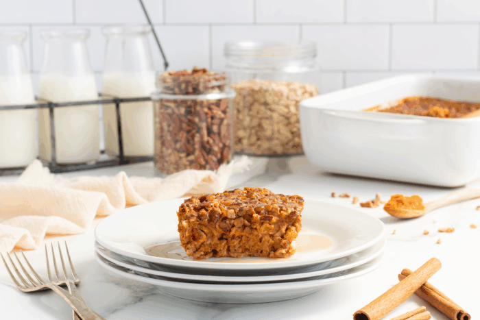 A slice of baked oatmeal sits on stacked white plates with syrup, surrounded by forks, cinnamon sticks, and jars of pecans, oats, and milk against a white tile background.