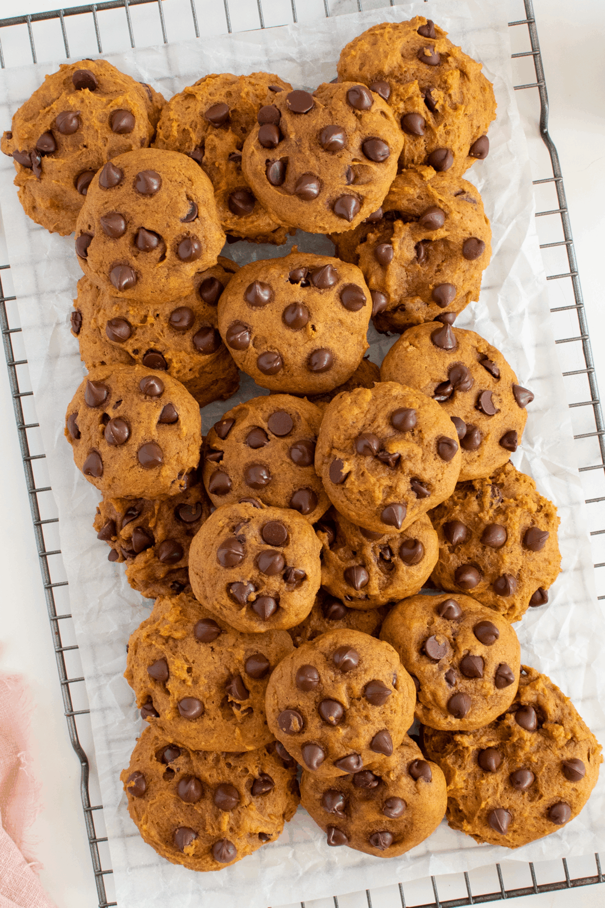 A batch of round, golden-brown cookies studded with chocolate chips is arranged on a wire cooling rack lined with white parchment paper. The cookies appear soft and freshly baked.