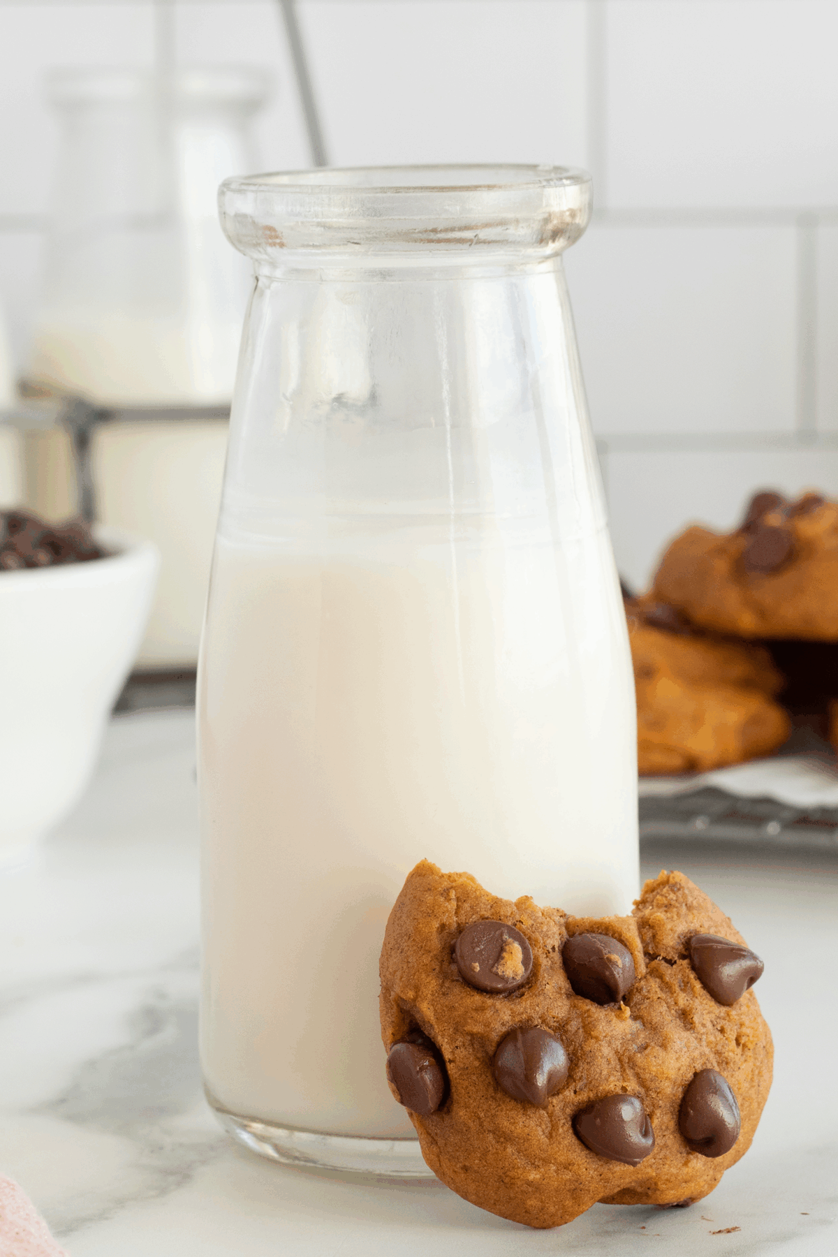 A glass bottle of milk with a chocolate chip cookie, partially bitten, leaning against it. In the background, more cookies and a bowl of chocolate chips are visible on a white surface.