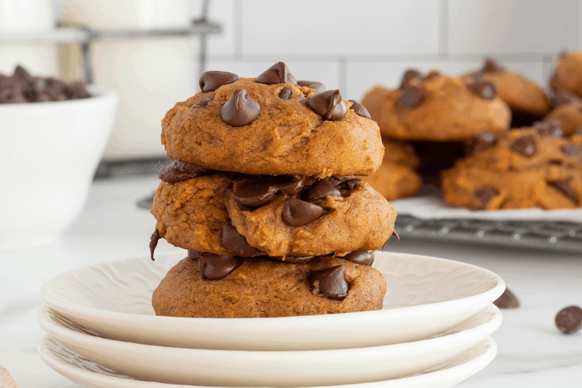 A stack of three chocolate chip cookies on a white plate, with more cookies and a bowl of chocolate chips in the background.