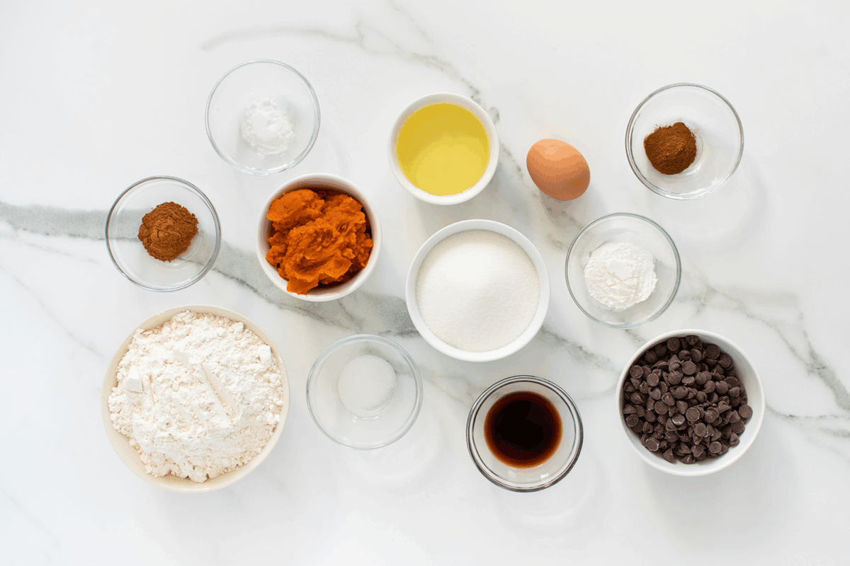 Top-down view of various baking ingredients in small glass bowls on a white marble surface, including flour, pumpkin puree, chocolate chips, egg, sugar, oil, vanilla, and spices.