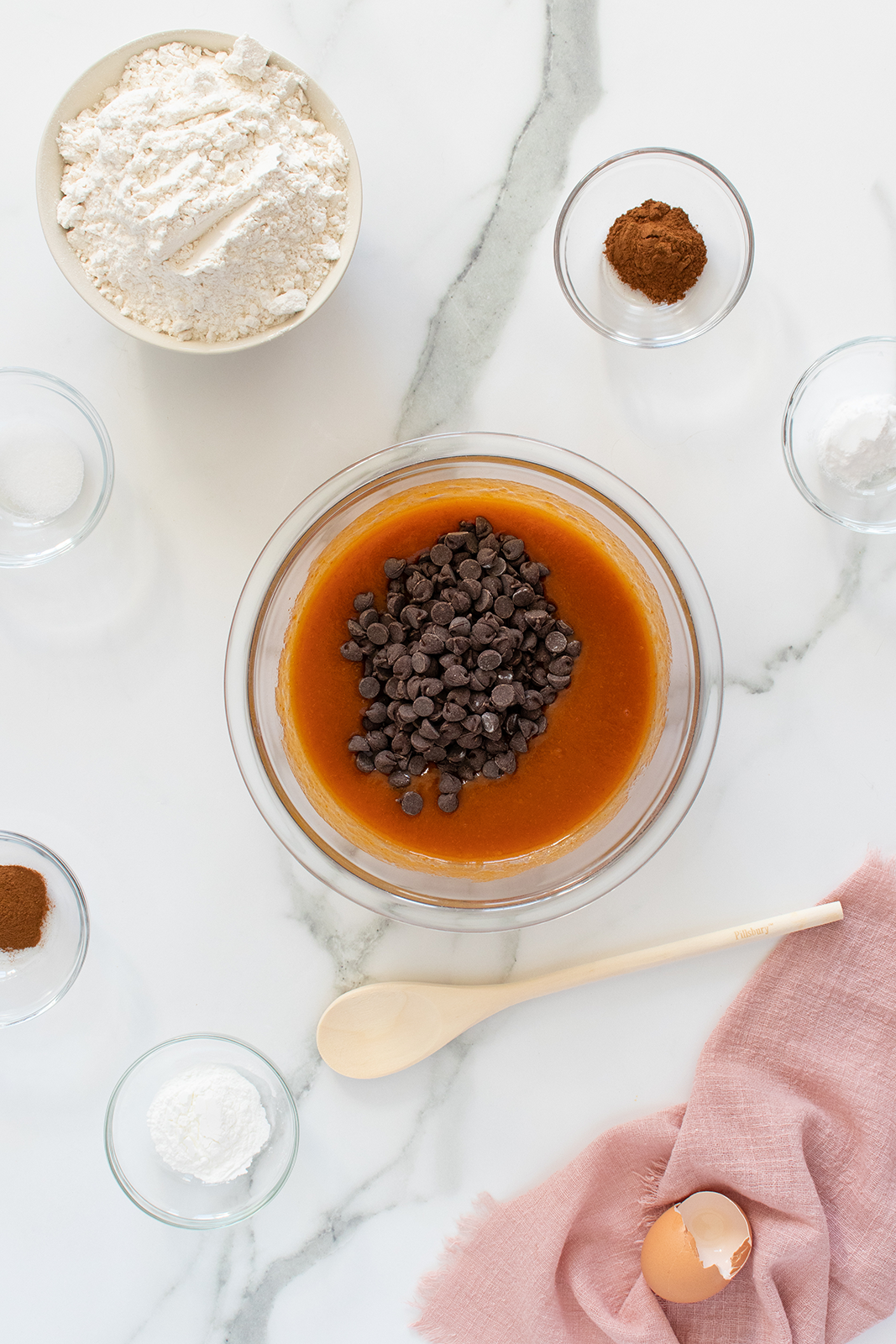 A mixing bowl with brown batter and chocolate chips, surrounded by bowls of flour, sugar, spices, cornstarch, and a cracked egg on a pink cloth, all on a white marble surface with a wooden spoon.