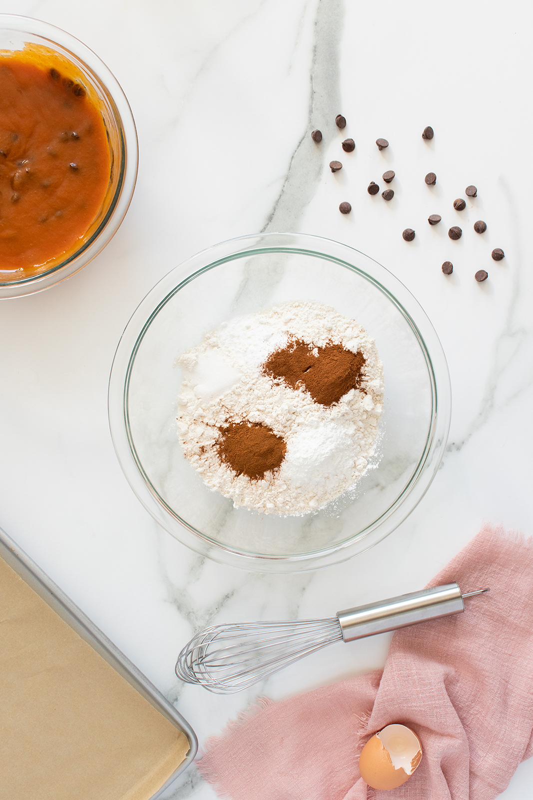 A glass bowl with flour, baking powder, and cinnamon on a marble surface, next to a whisk, broken eggshell, pink cloth, scattered chocolate chips, baking pan, and a bowl of pumpkin mixture.