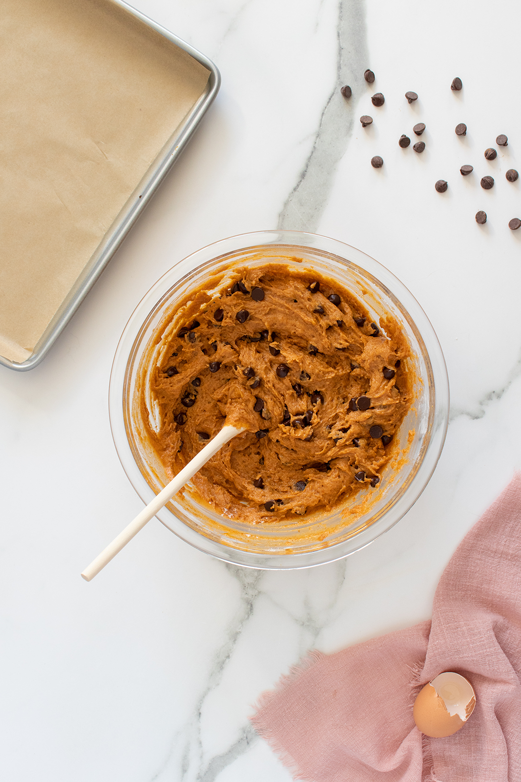 A glass bowl filled with cookie dough and chocolate chips sits on a marble countertop. A wooden spoon rests in the bowl. Nearby are a baking sheet with parchment, scattered chocolate chips, a pink cloth, and a cracked eggshell.
