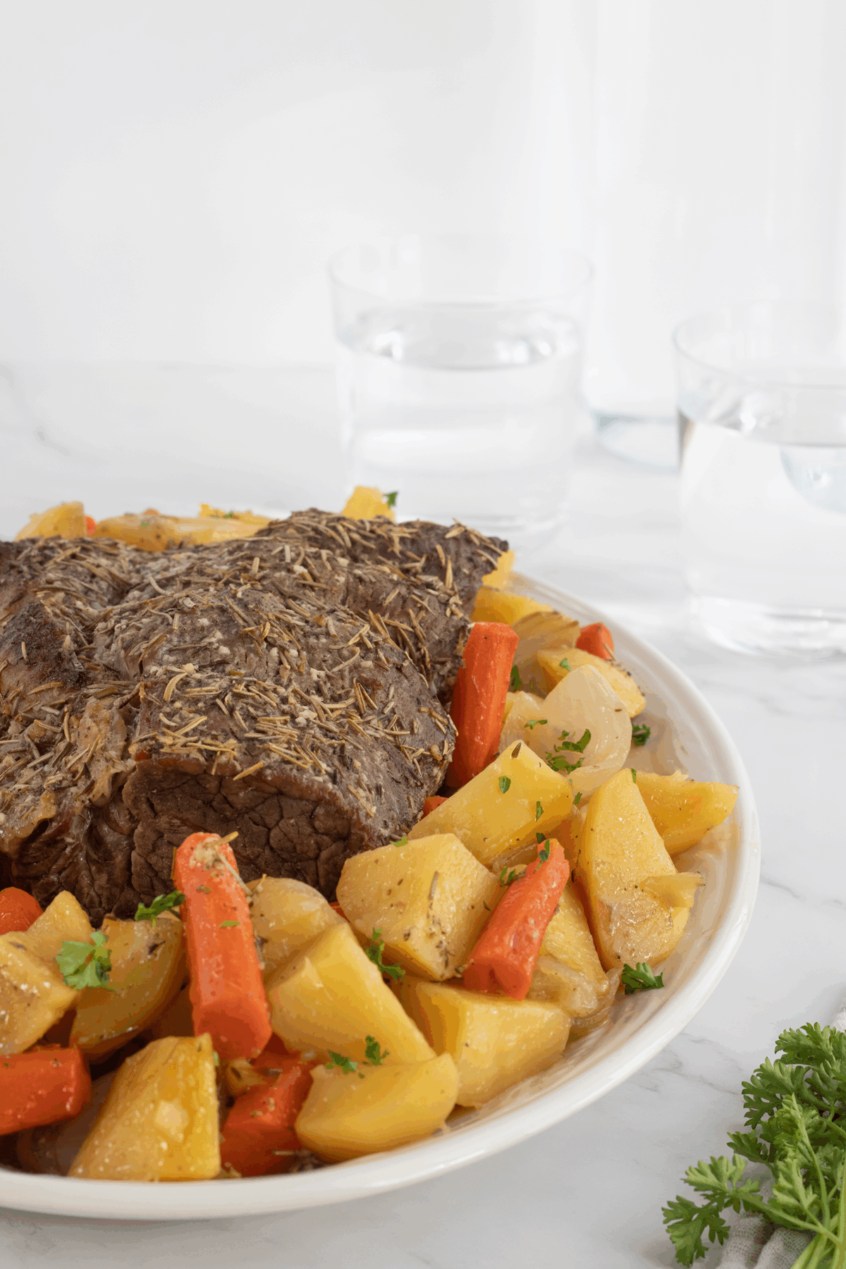 A plate with pot roast topped with herbs, surrounded by cooked carrots, potatoes, and onions. Two glasses of water in the background and a sprig of parsley on the side, all set on a white surface.