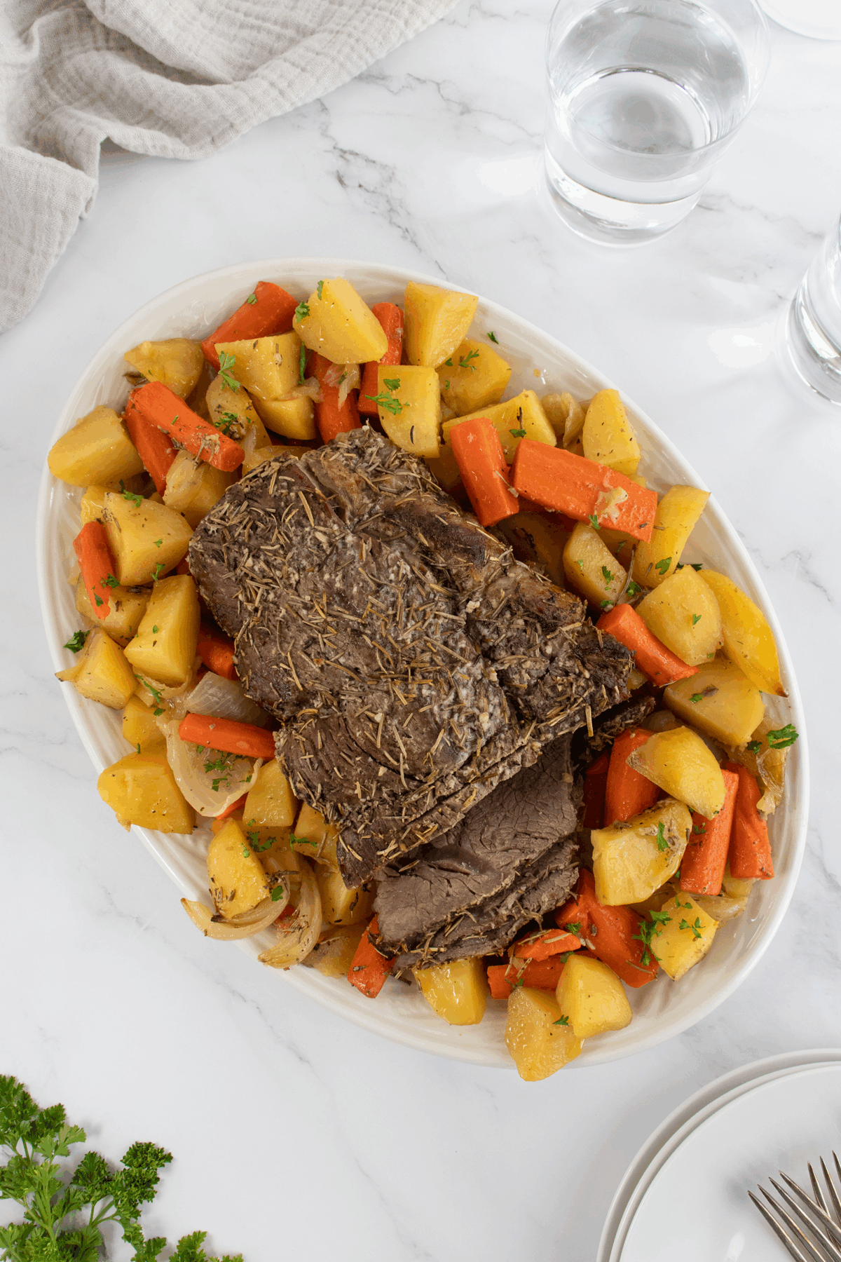 An oval platter with sliced roast beef garnished with herbs, surrounded by cooked potatoes and carrots, sits on a marble surface next to two glasses of water and a beige napkin.