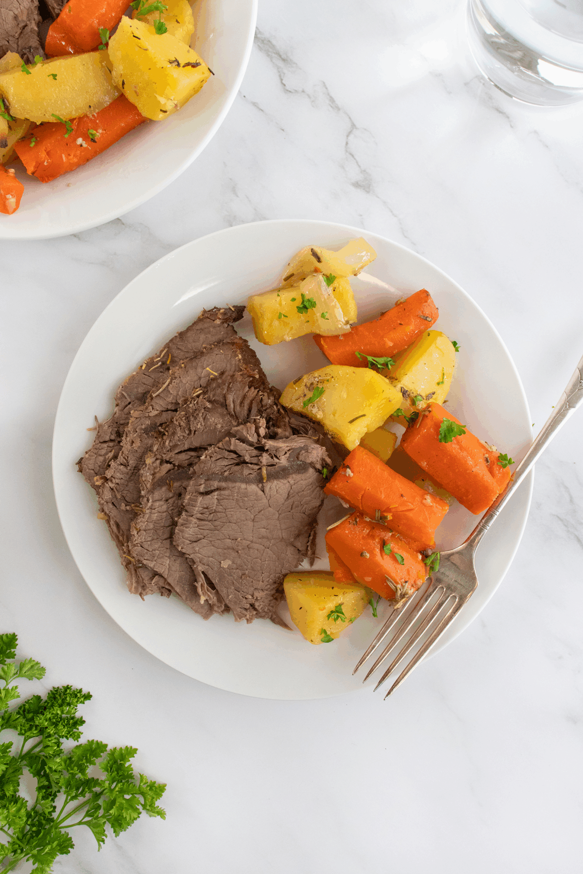 A white plate with sliced pot roast, cooked carrots, and potatoes garnished with parsley, next to a fork. Another plate and a glass of water are partially visible in the background on a marble surface.